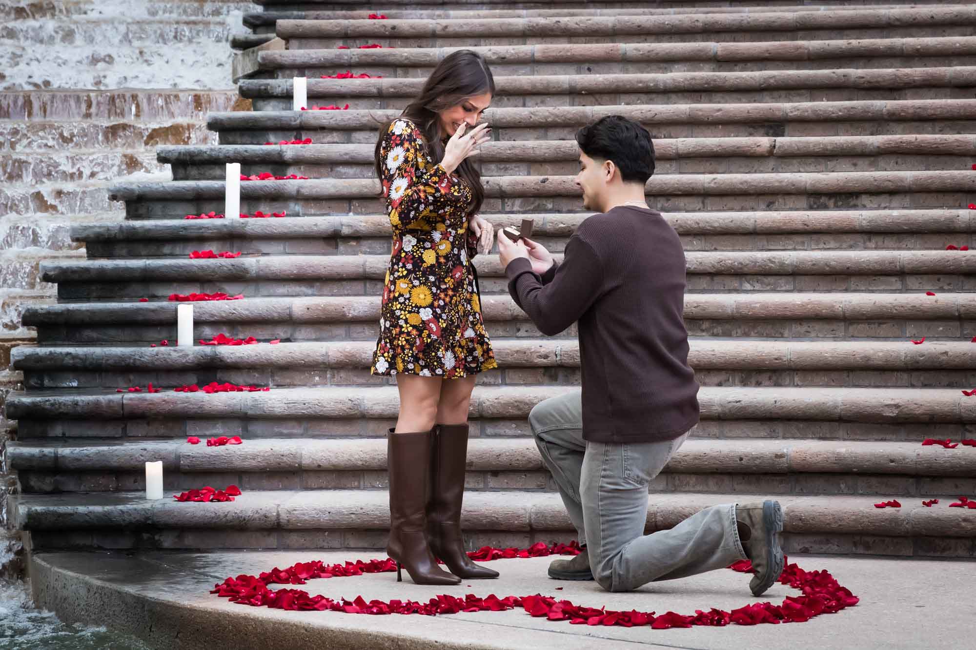 Man proposing to woman on bended knee at the base of the stairs of the Weston Centre in a heart-shaped ring of rose petals during a downtown San Antonio surprise proposal
