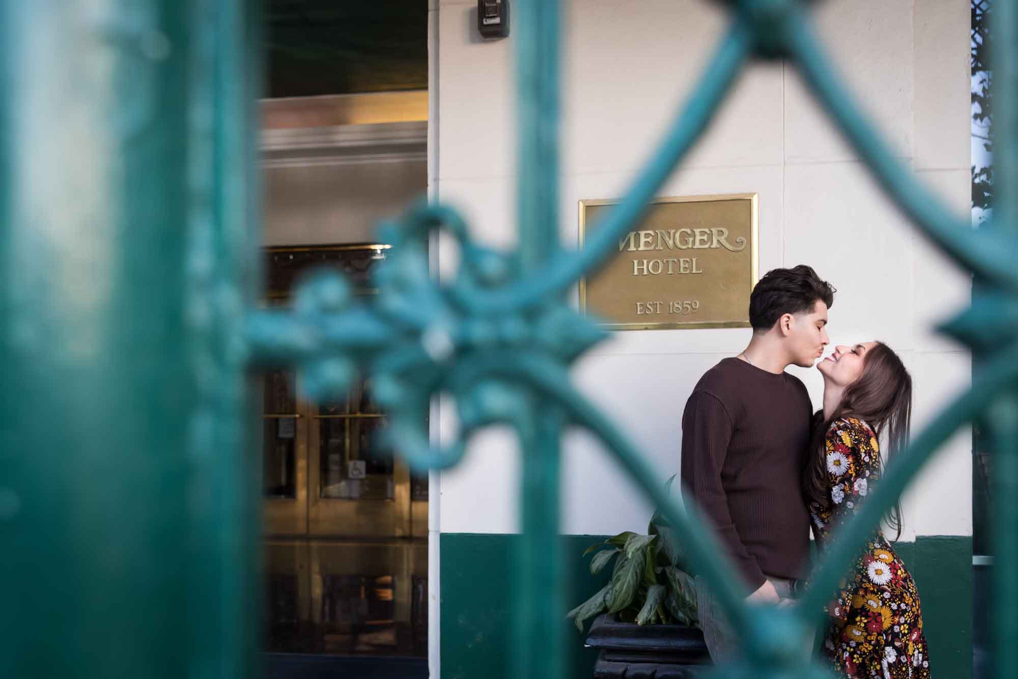 View of couple kissing through green wrought iron gate of Menger Hotel during a downtown San Antonio surprise proposal