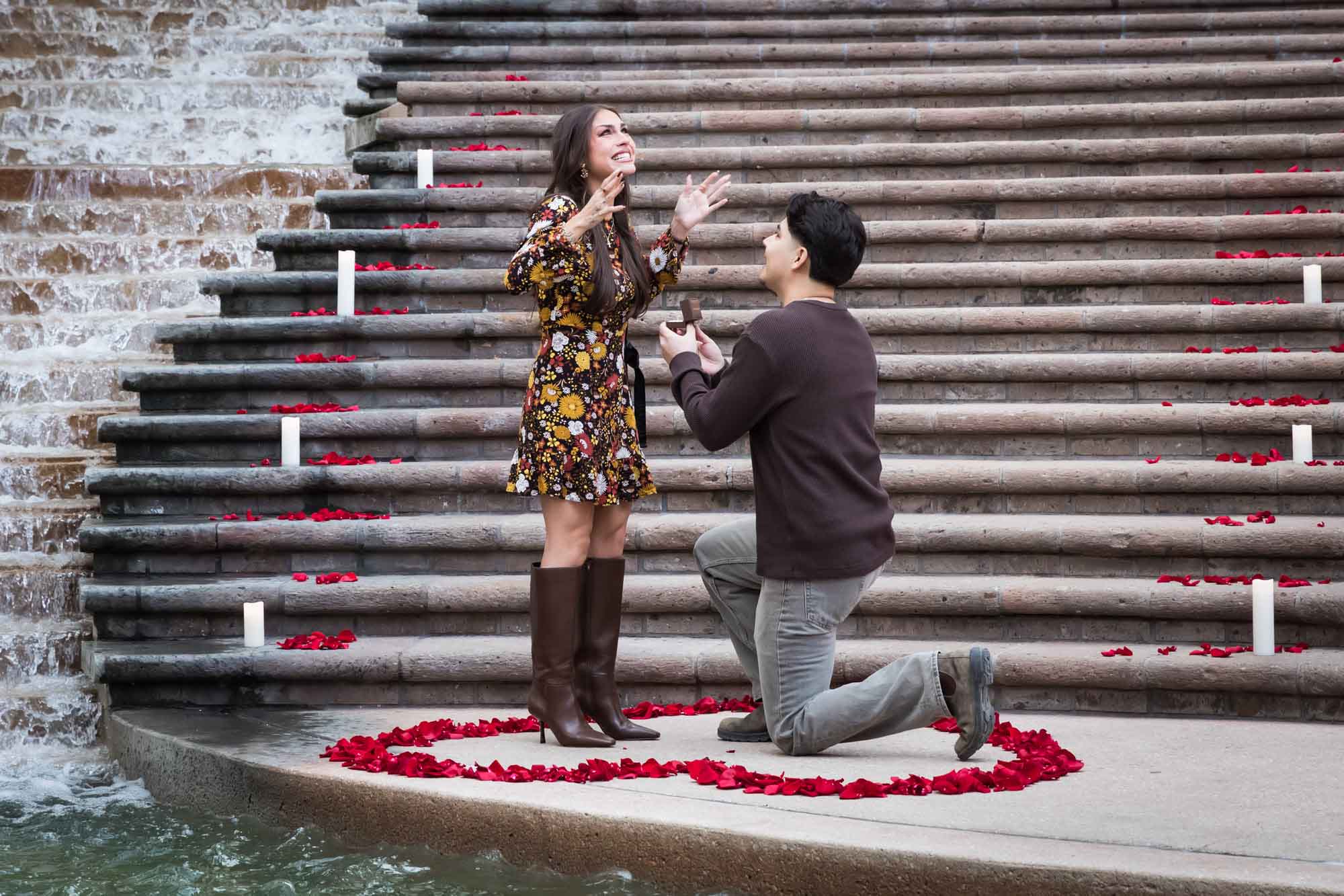 Man proposing to woman on bended knee at the base of the stairs of the Weston Centre in a heart-shaped ring of rose petals during a downtown San Antonio surprise proposal