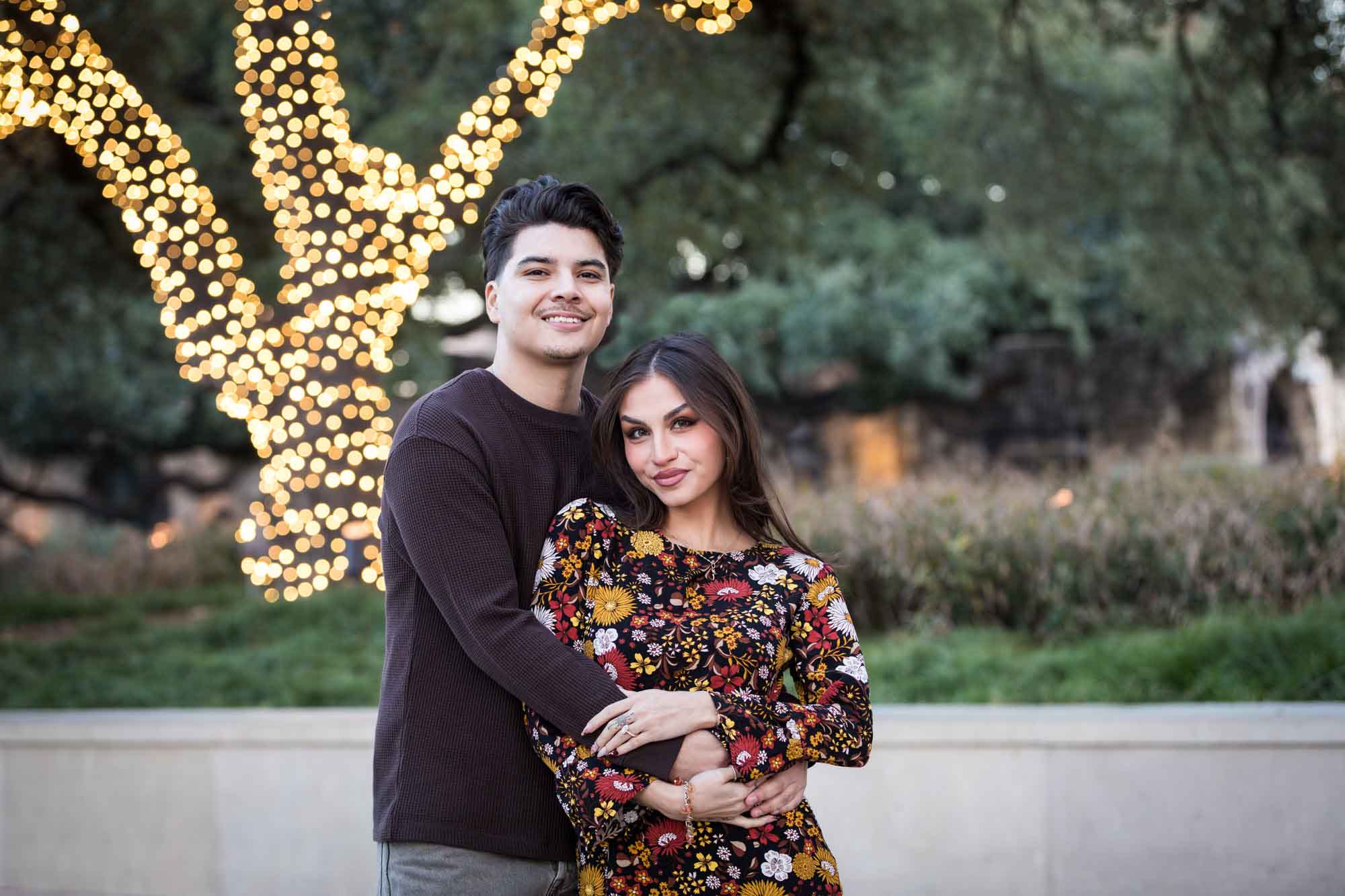 Man and woman hugging in front of brightly lit tree during a downtown San Antonio surprise proposal