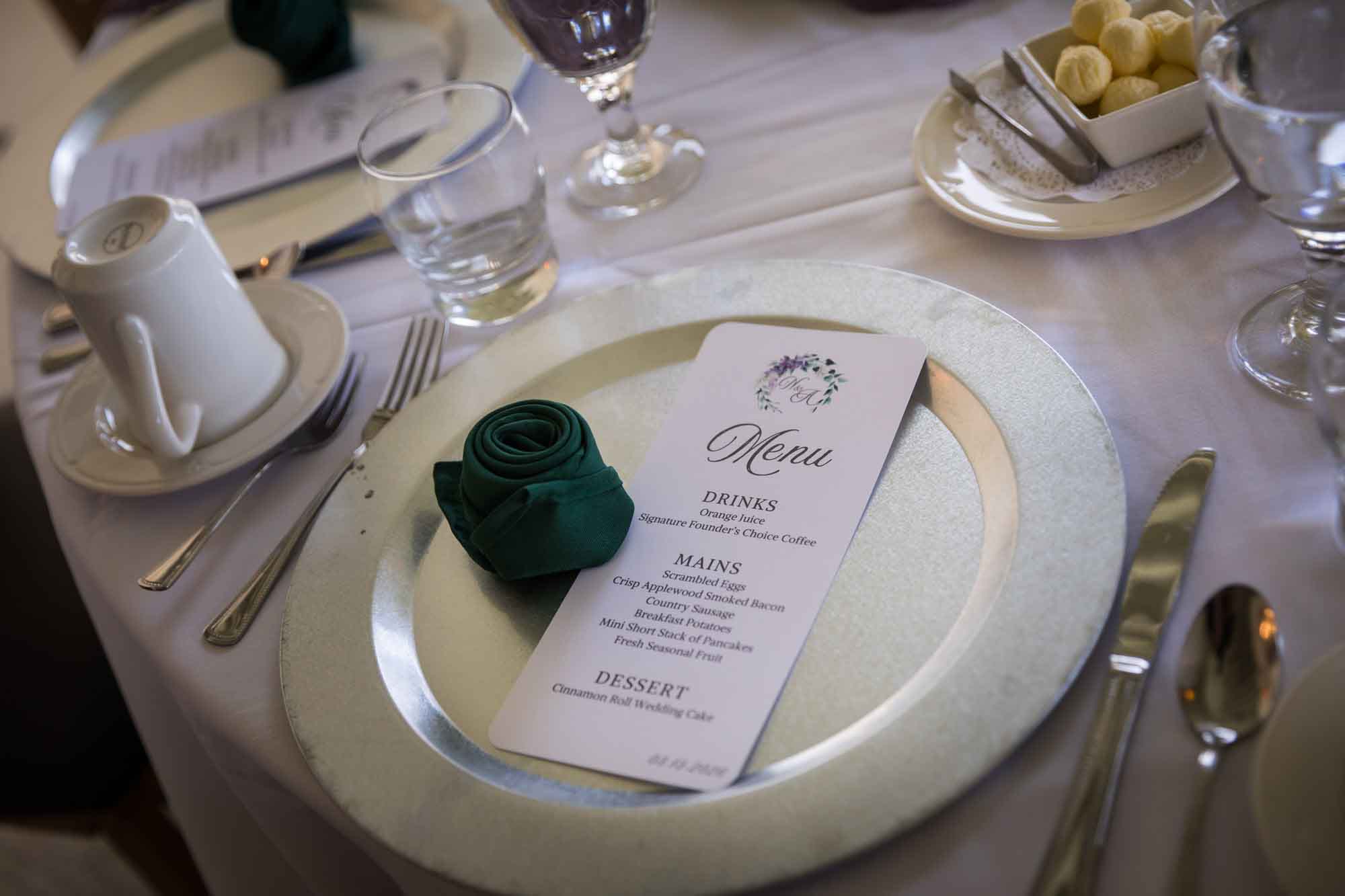 Place setting of gold charger set with green, wrapped napkin, menu card, and silverware during a Guenther House wedding reception