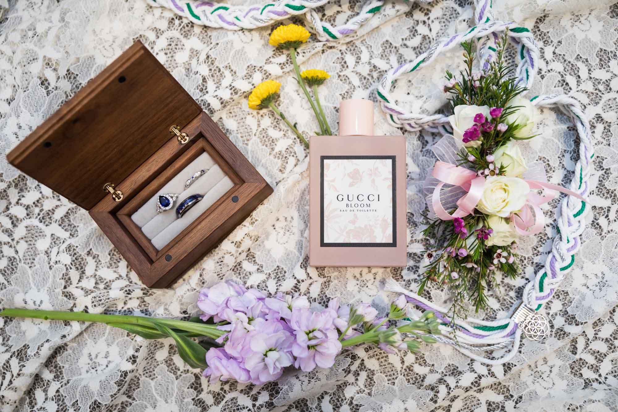 Display of flowers, Gucci perfume bottle, wooden box with rings, and woven rope on lace fabric before a San Antonio Botanical Garden wedding ceremony