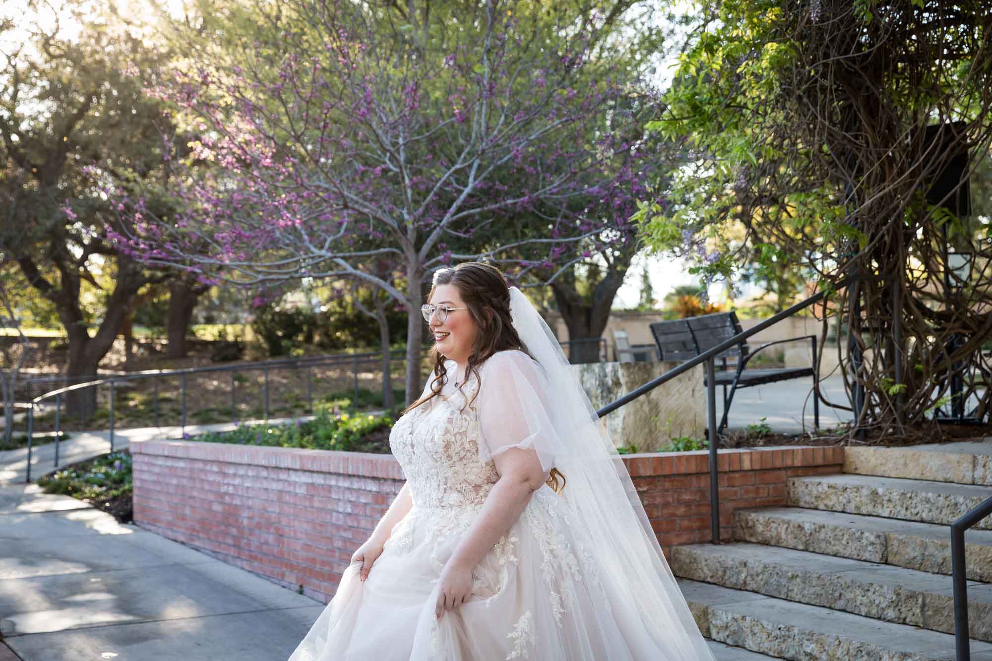 Bride wearing white dress and long veil standing in front of stairs and purple tree before a San Antonio Botanical Garden wedding ceremony