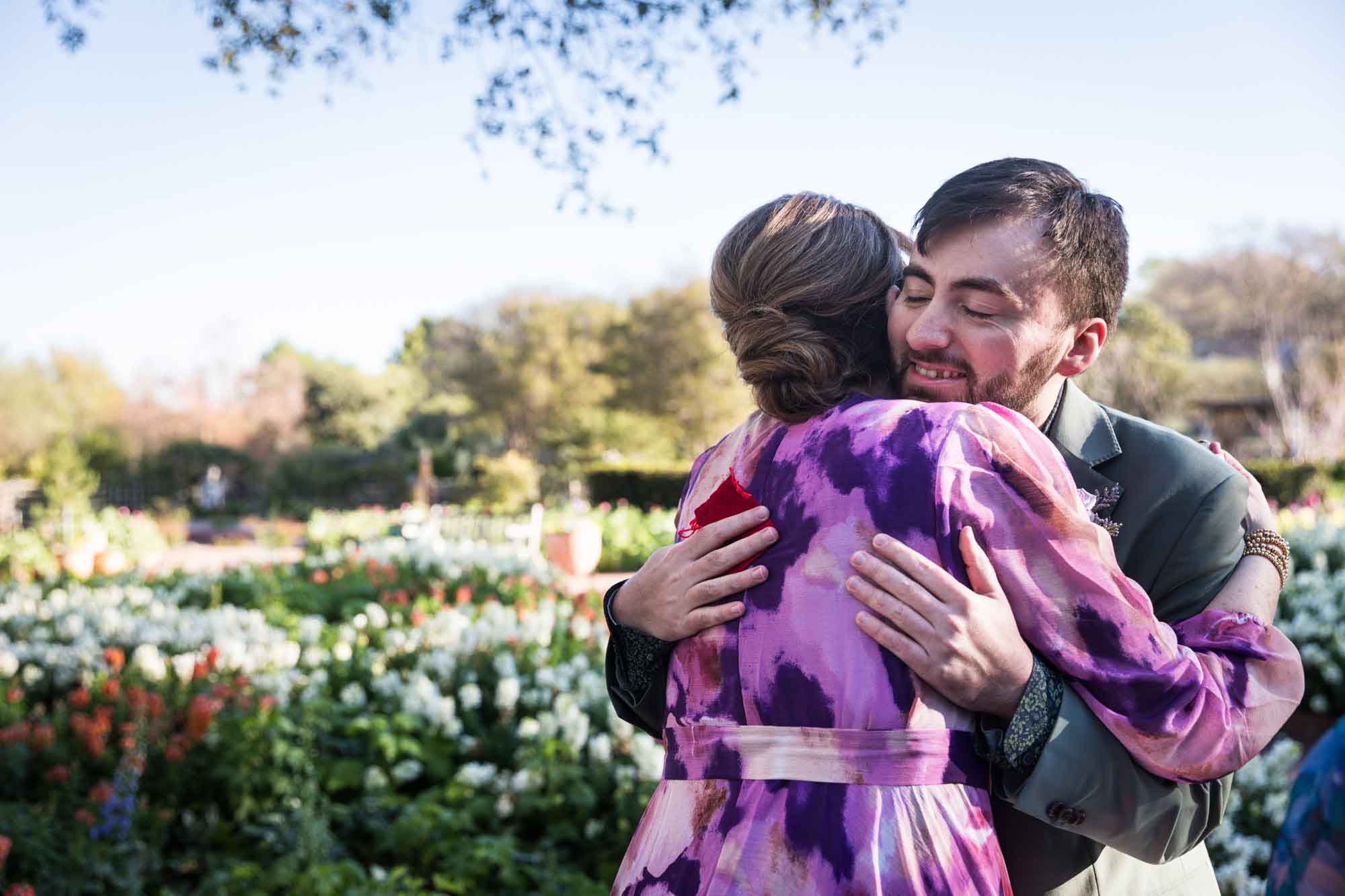 Groom hugging woman wearing purple floral dress in front of flower garden before a San Antonio Botanical Garden wedding ceremony
