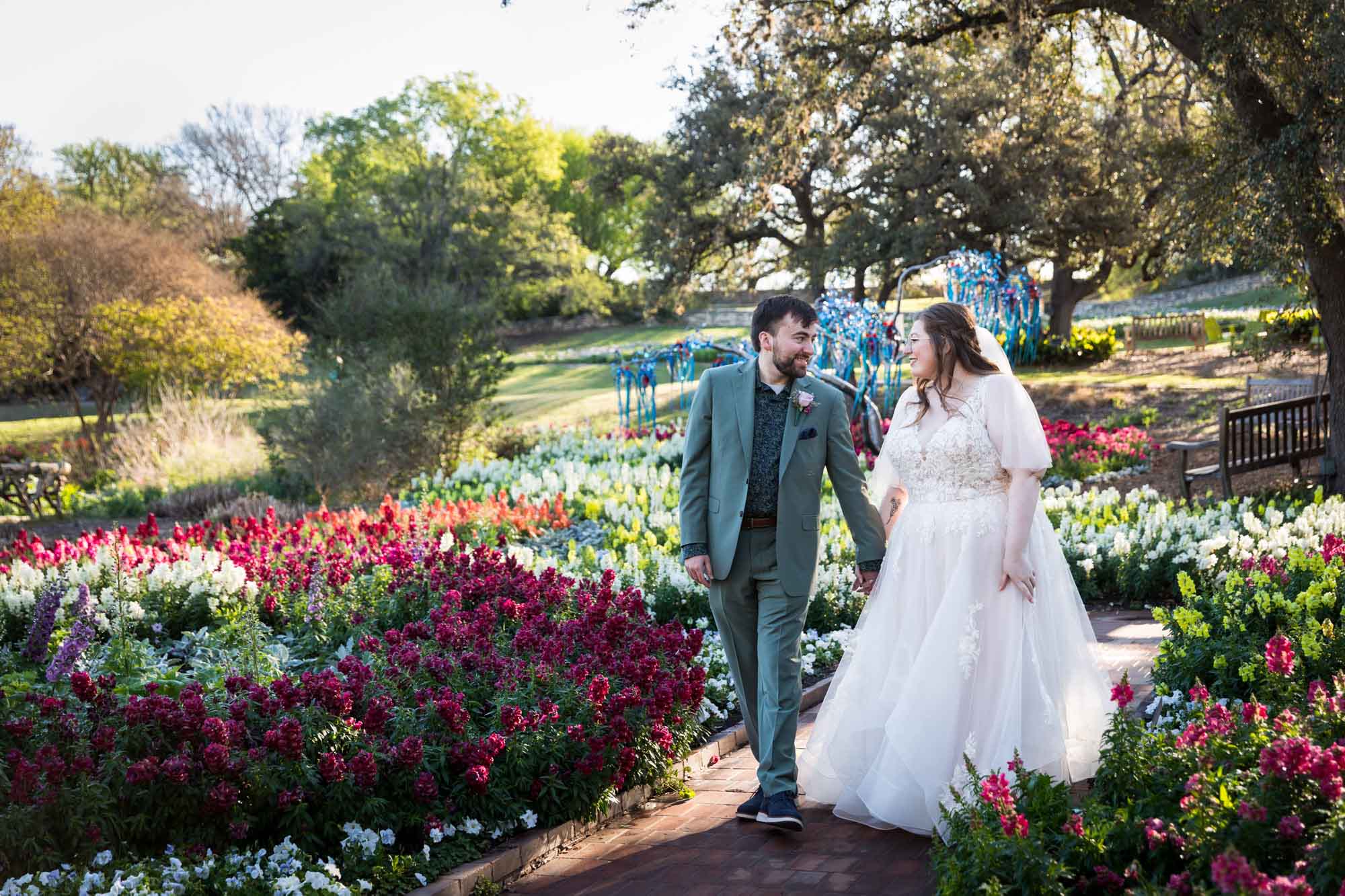 Bride and groom holding hands walking in front of bed of colorful snapdragons and trees before a San Antonio Botanical Garden wedding ceremony