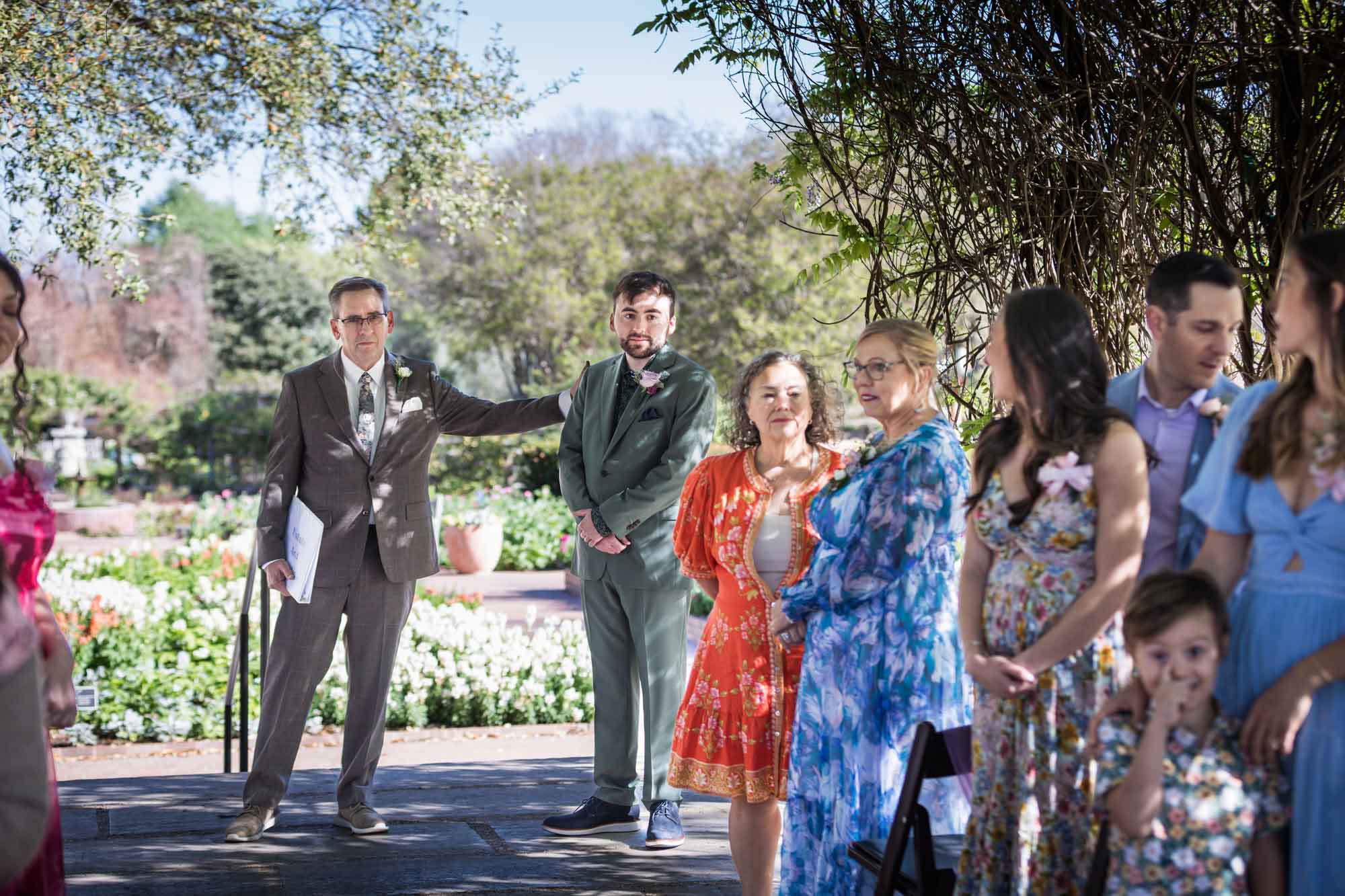 Father with hand on groom's shoulder standing behind guests during a San Antonio Botanical Garden wedding ceremony