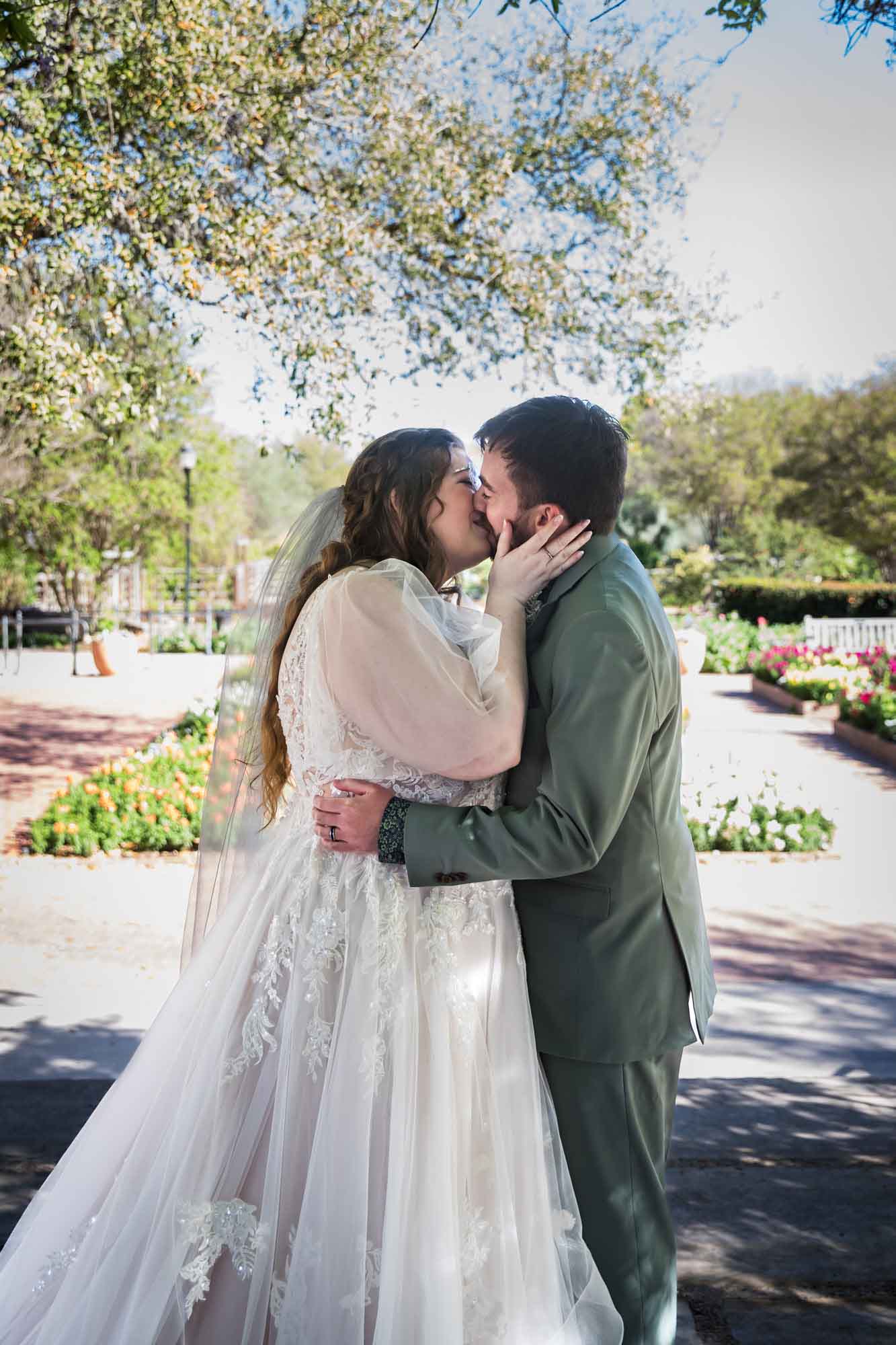 Bride and groom kissing with bride's hand on groom's face in front of colorful flowers during a San Antonio Botanical Garden wedding ceremony