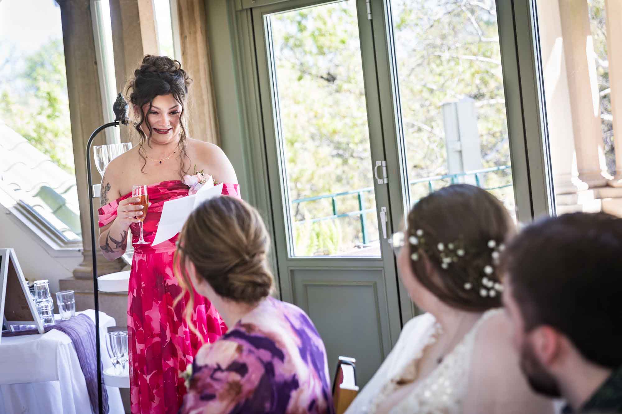 Woman wearing red, floral dress holding paper and glass of champagne in front of guests and glass door during speech during a Guenther House wedding reception