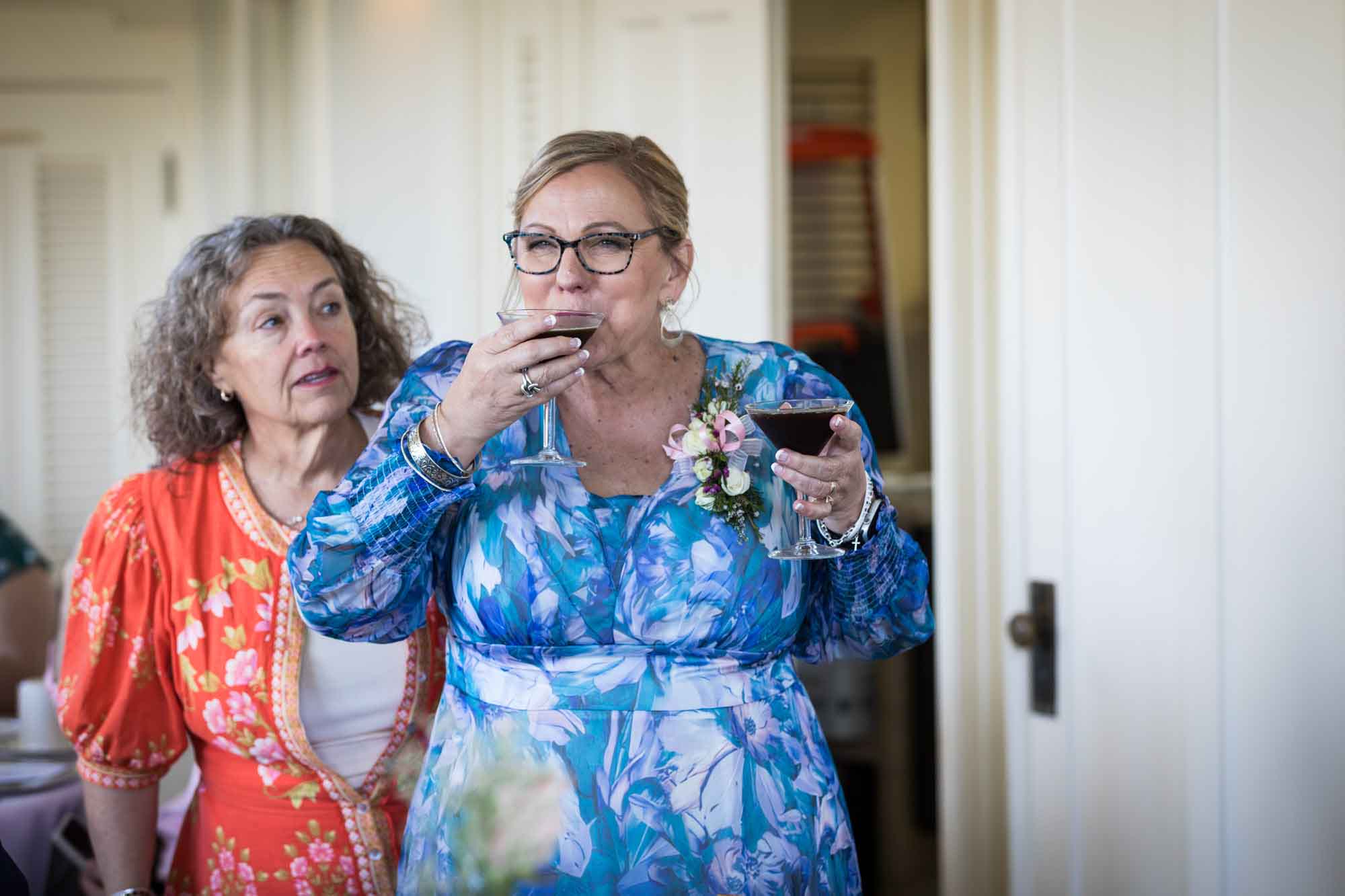 Woman wearing blue floral dress holding and drinking two espresso martinis in front of woman wearing orange dress during a Guenther House wedding reception