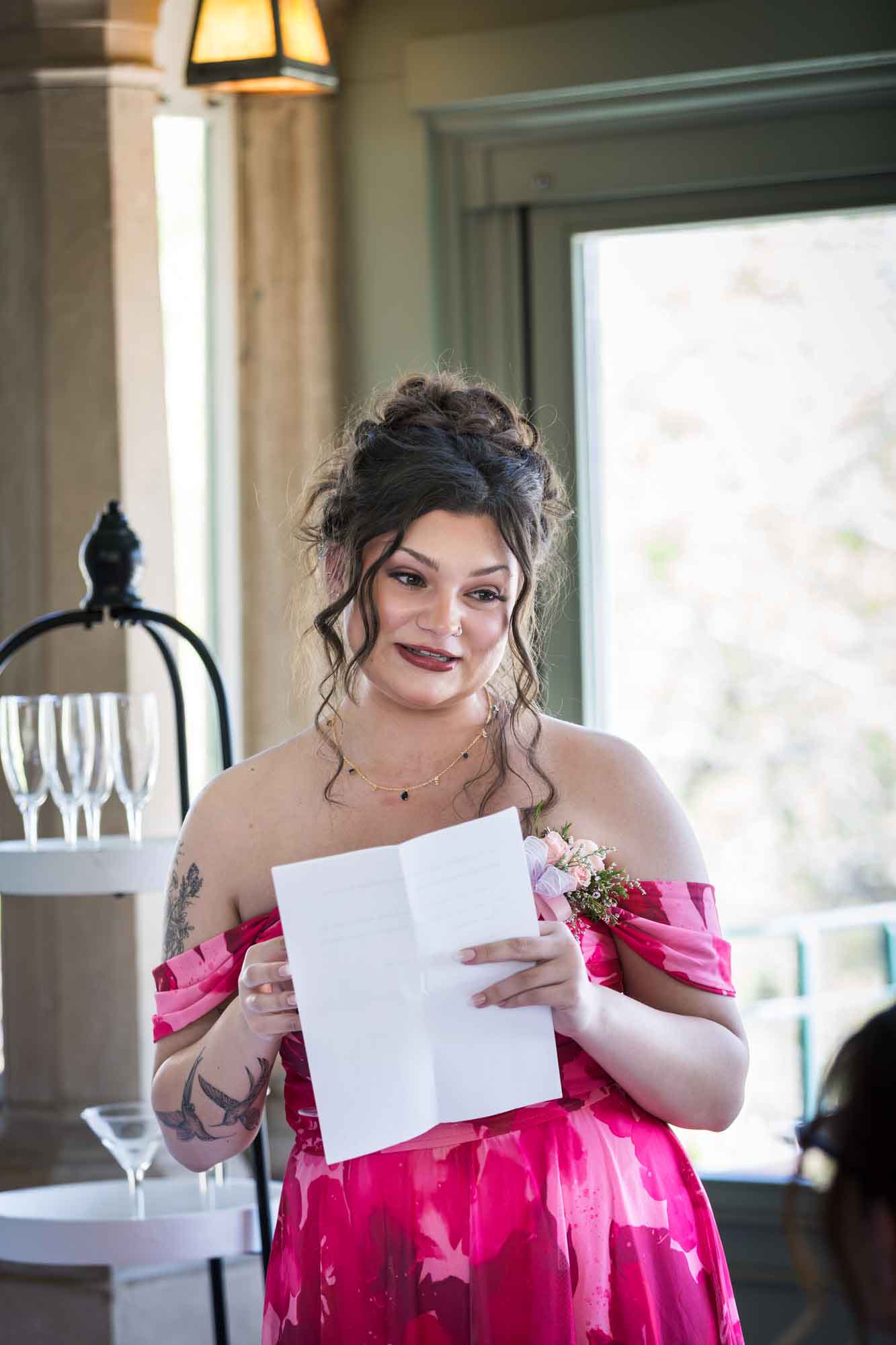Woman wearing red, sleeveless floral dress and holding white piece of paper in front of window during speech during a Guenther House wedding reception