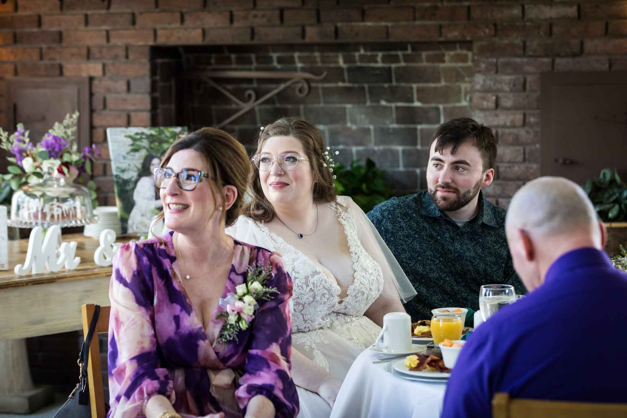 Bride and groom listening to speech while seated in front of guests and brick fireplace during a Guenther House wedding reception