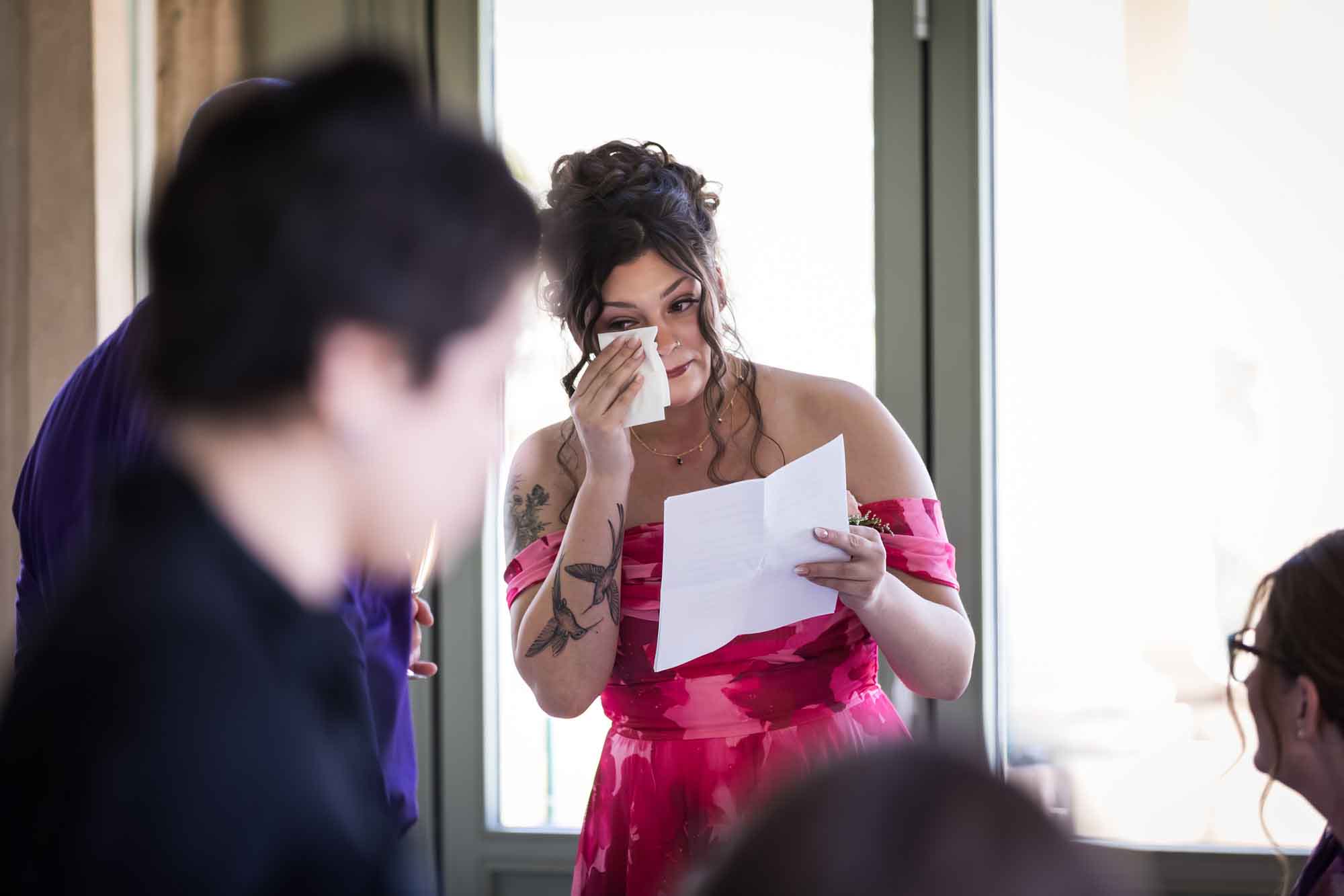 Woman wearing red floral dress holding paper and wiping tears with tissue during a Guenther House wedding reception