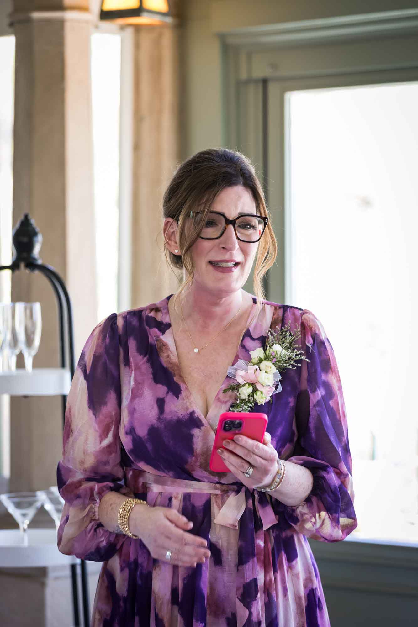 Woman wearing purple floral dress holding pink cell phone in front of door during speech during a Guenther House wedding reception