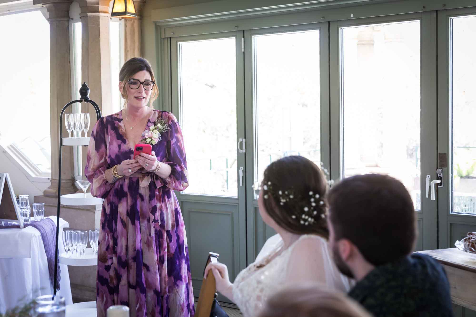 Woman wearing purple floral dress holding pink cell phone speaking to bride and groom during speech during a Guenther House wedding reception