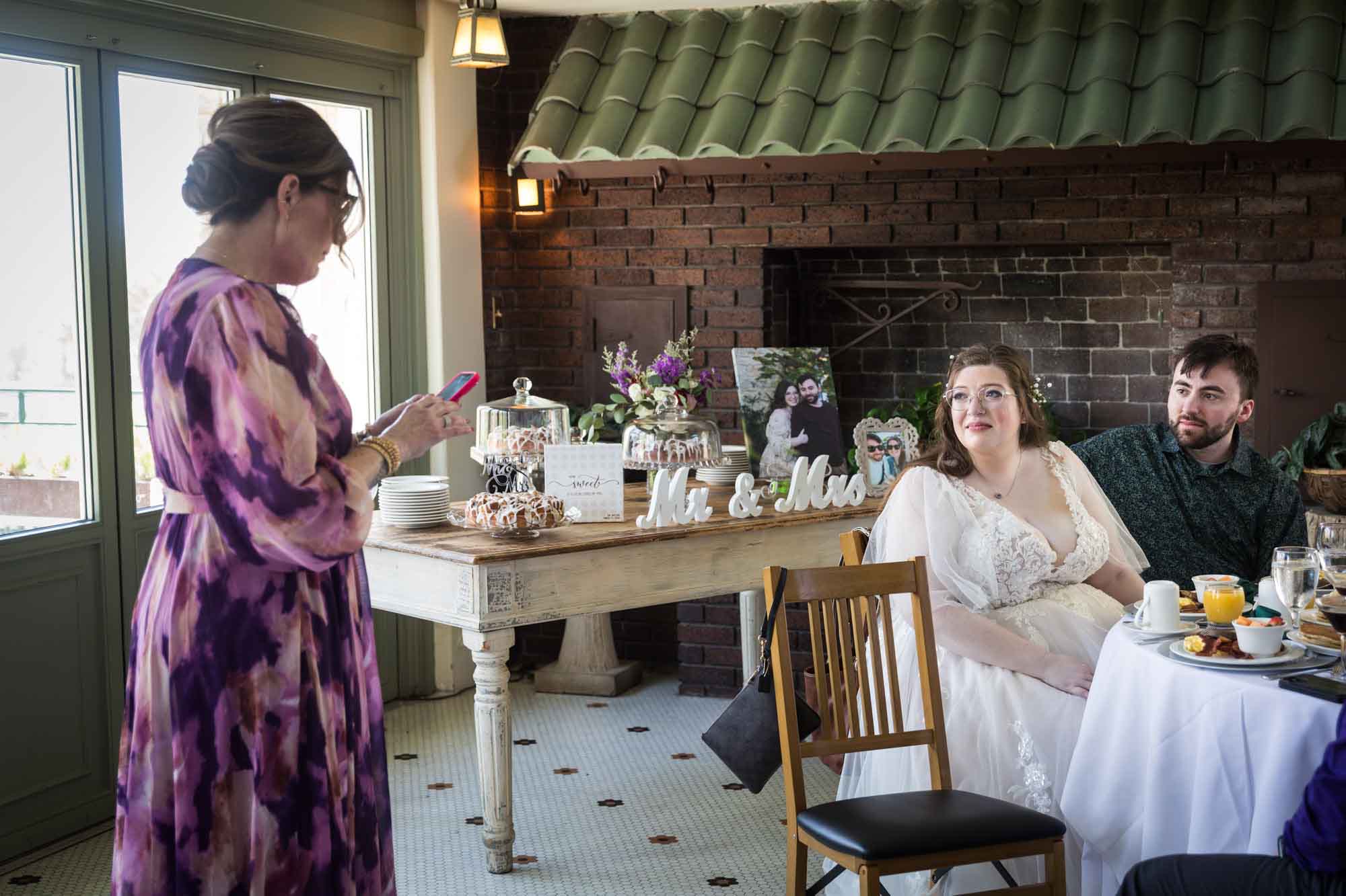 Bride and groom seated listening to woman wearing purple floral dress reading from cell phone during speech during a Guenther House wedding reception