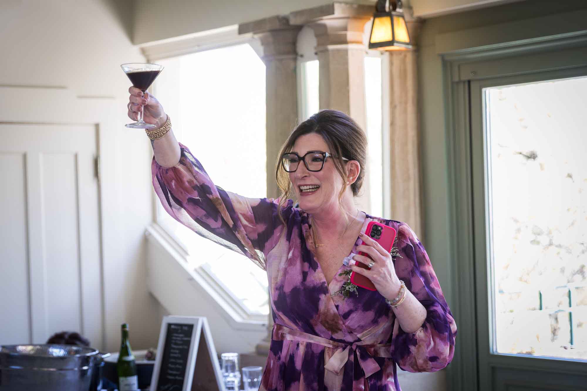 Woman wearing purple floral dress holding pink cell phone and espresso martini in the air during a Guenther House wedding reception