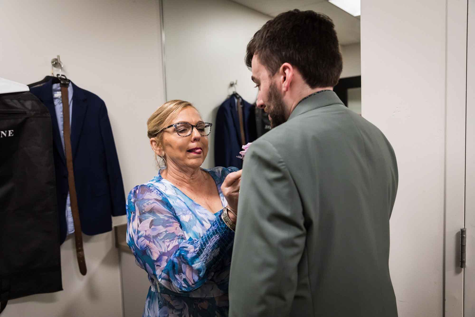 Mother putting flower on lapel of groom while holding out tongue in room before a San Antonio Botanical Garden wedding ceremony