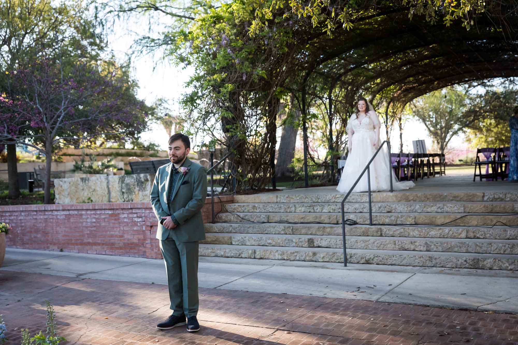 Groom with back turned to bride who is standing on stairs under wisteria arbor looking at groom before a San Antonio Botanical Garden wedding ceremony
