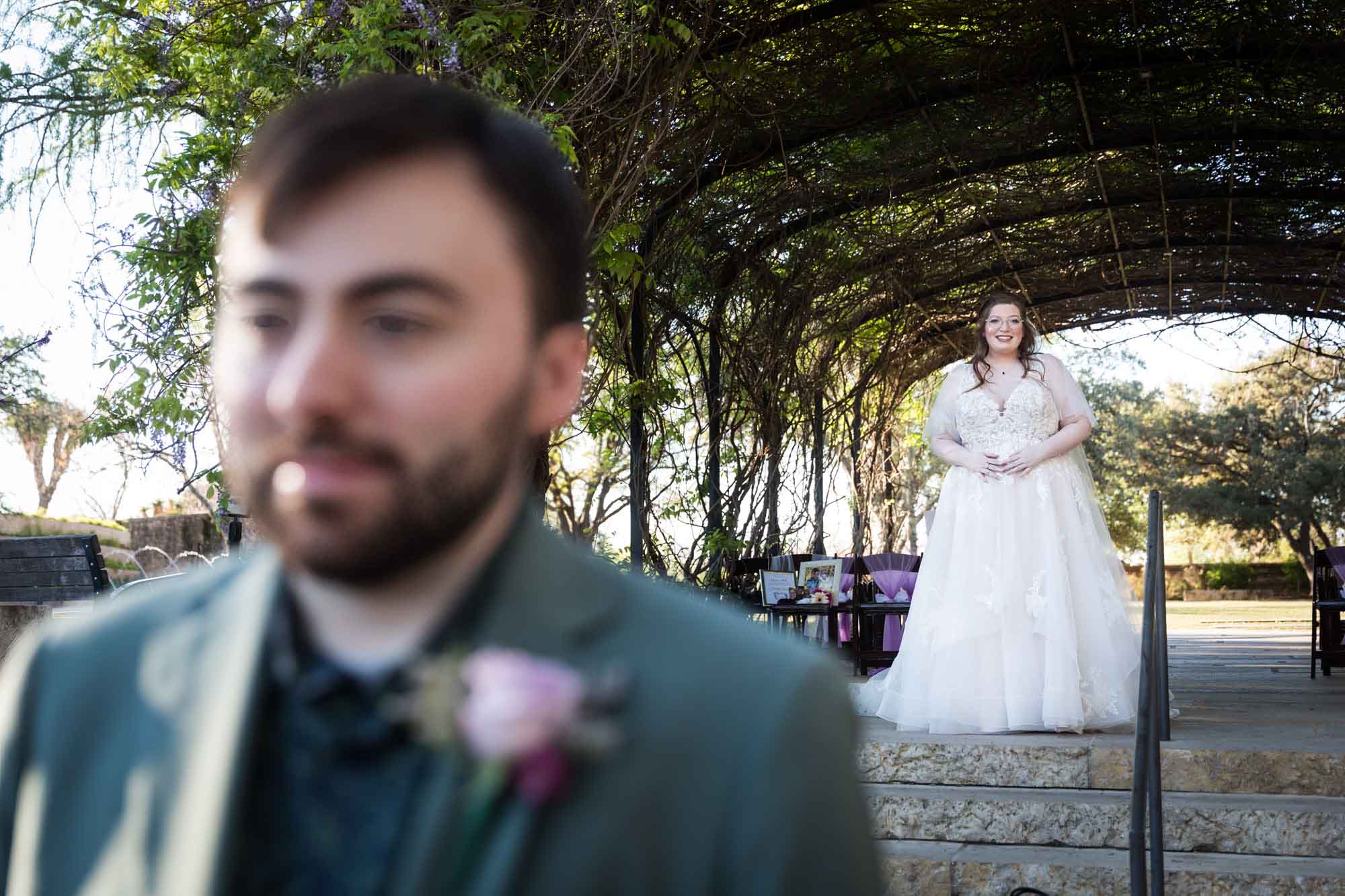 Bride standing on stone stairs under Wisteria ARbor looking down at groom before a San Antonio Botanical Garden wedding ceremony