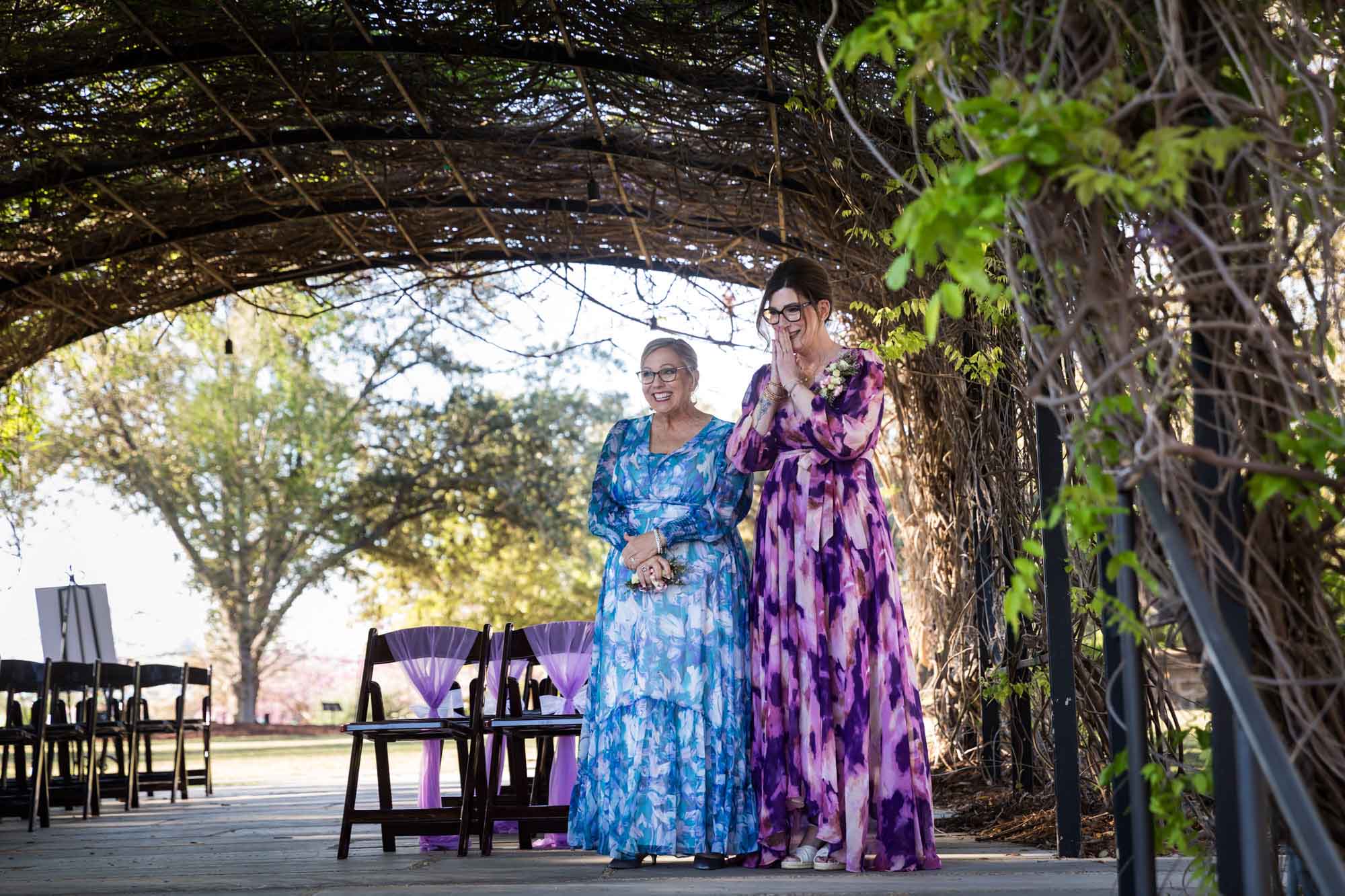 Two women wearing floral dresses under Wisteria Arbor standing in front of chairs before a San Antonio Botanical Garden wedding ceremony