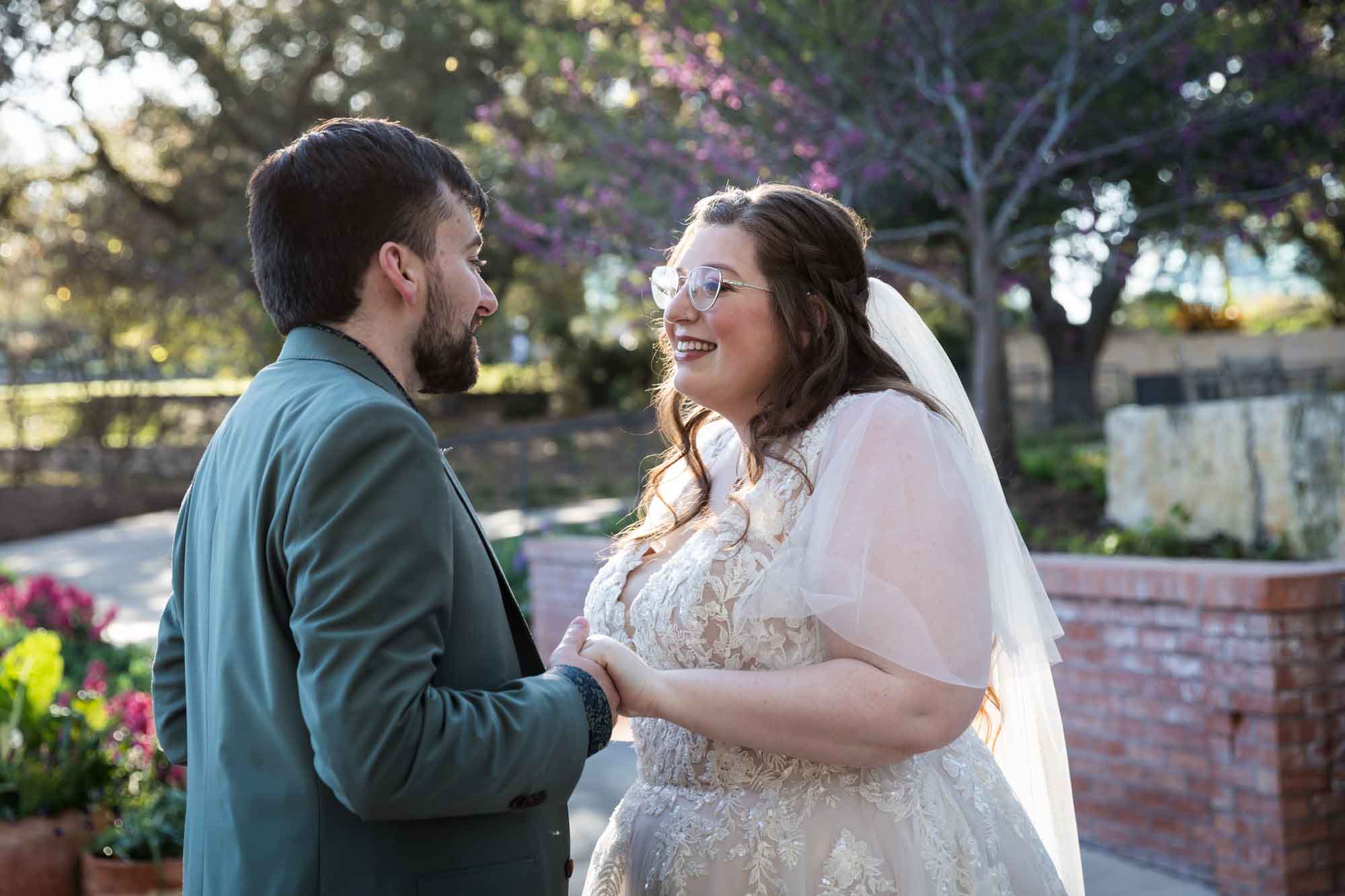 Bride wearing white dress and veil holding hands with groom wearing green blazer in front of flowers and brick wall before a San Antonio Botanical Garden wedding ceremony