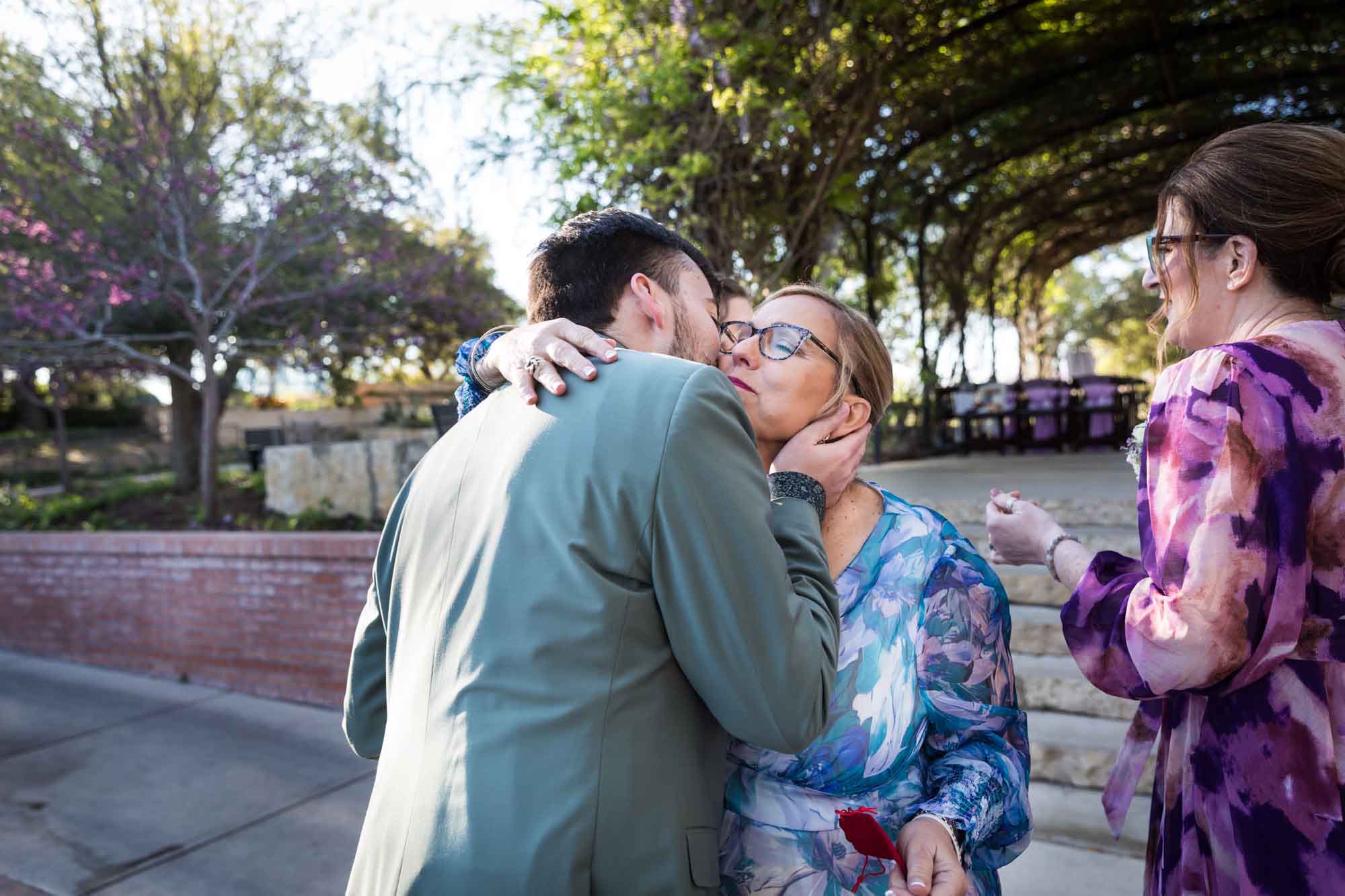 Woman wearing blue floral dress hugging man wearing green jacket before a San Antonio Botanical Garden wedding ceremony