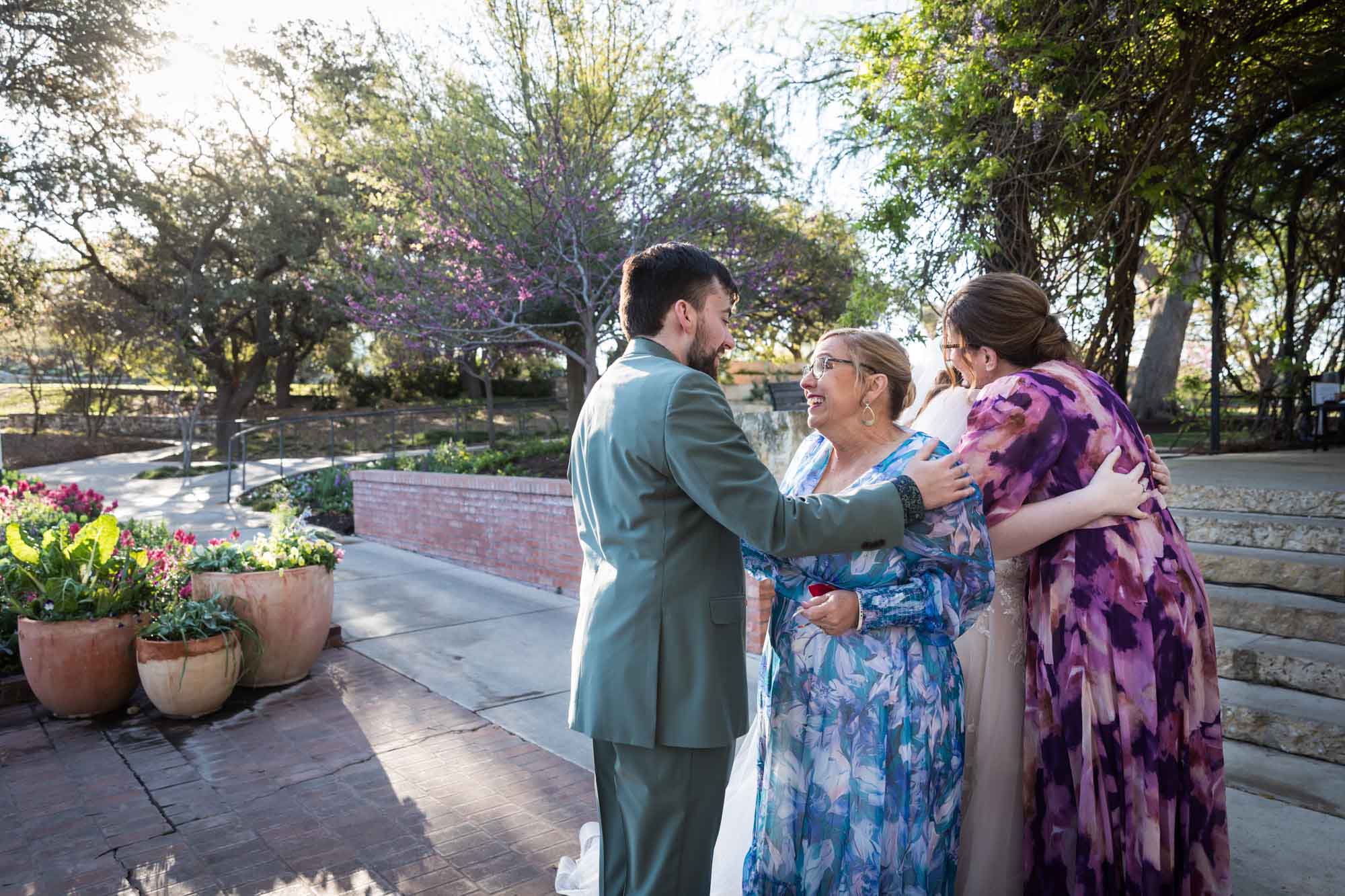 Two women wearing floral dresses hugging bride and groom in front of planters of flowers, stairs, and brick wall before a San Antonio Botanical Garden wedding ceremony