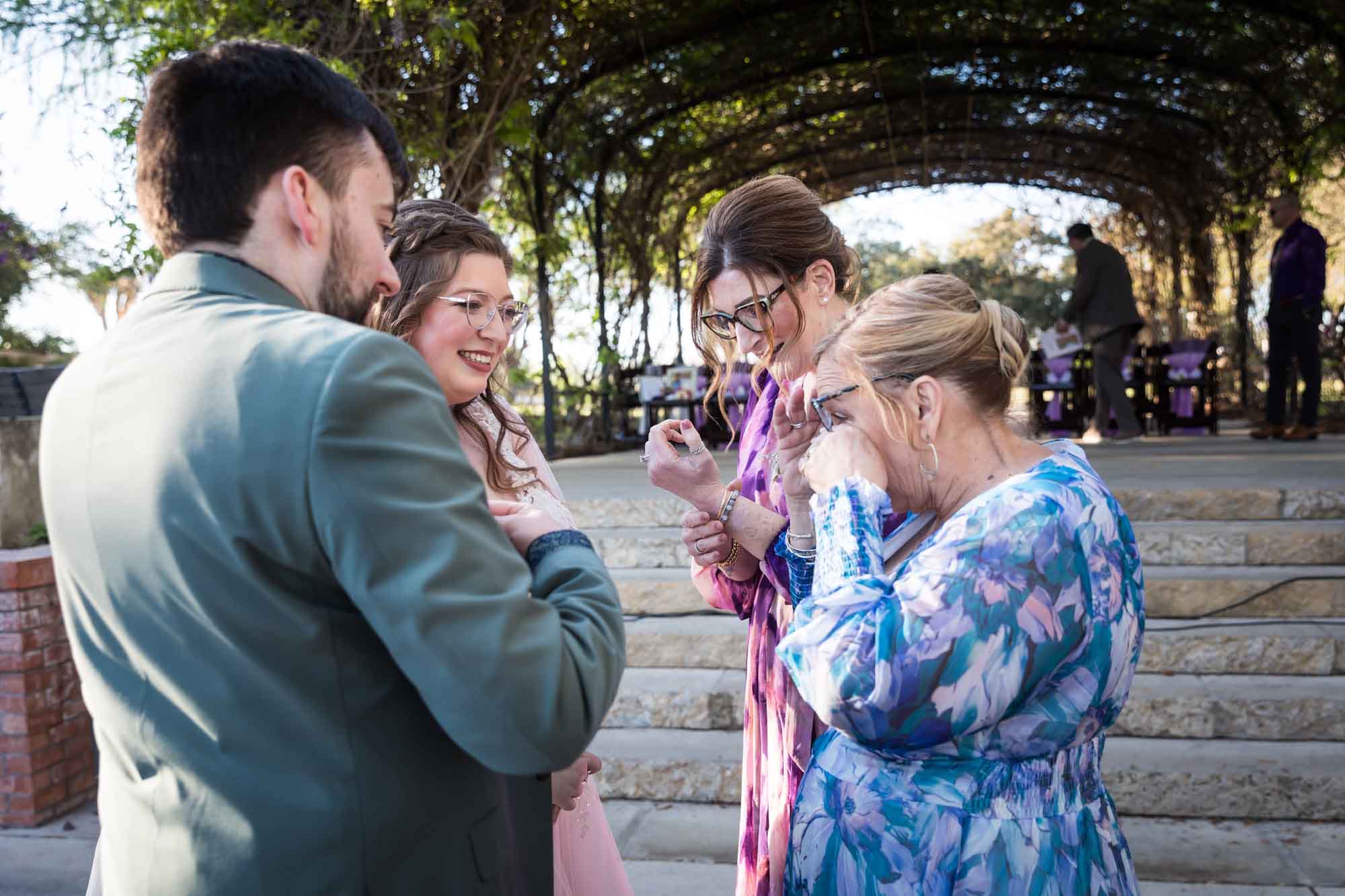Bride and groom looking over at mothers who are wiping tears and inspecting bracelet on wrist before a San Antonio Botanical Garden wedding ceremony