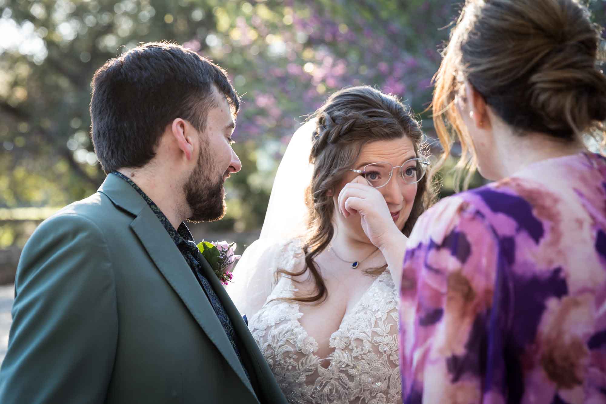 Bride wearing glasses wiping away tear standing beside groom wearing green jacket and woman wearing purple floral dress before a San Antonio Botanical Garden wedding ceremony