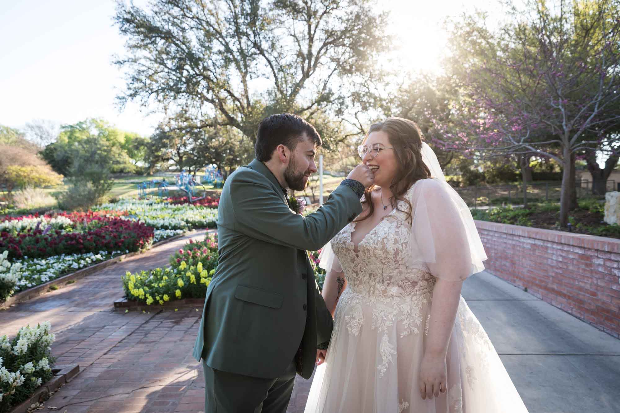 Groom wearing green jacket wiping something off teeth of bride wearing white dress in front of flower garden and brick wall before a San Antonio Botanical Garden wedding ceremony