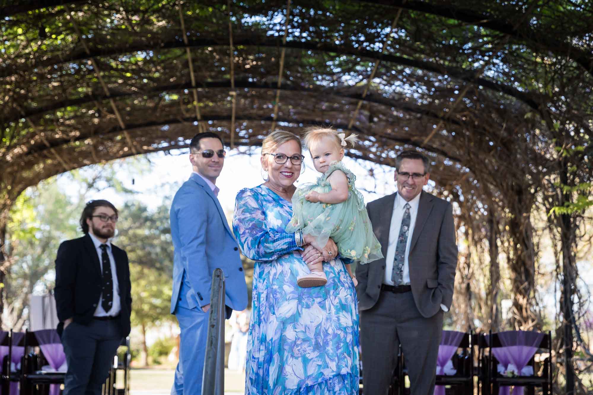 Woman wearing blue floral dress holding baby girl wearing green dress in front of three men under Wisteria Arbor before a San Antonio Botanical Garden wedding ceremony