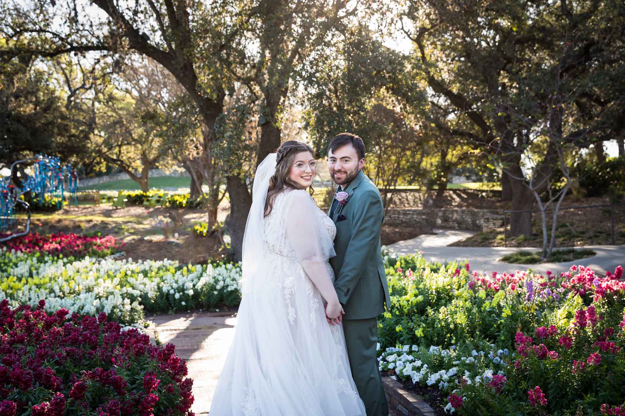 Bride and groom holding hands in front of bed of colorful snapdragons and trees before a San Antonio Botanical Garden wedding ceremony
