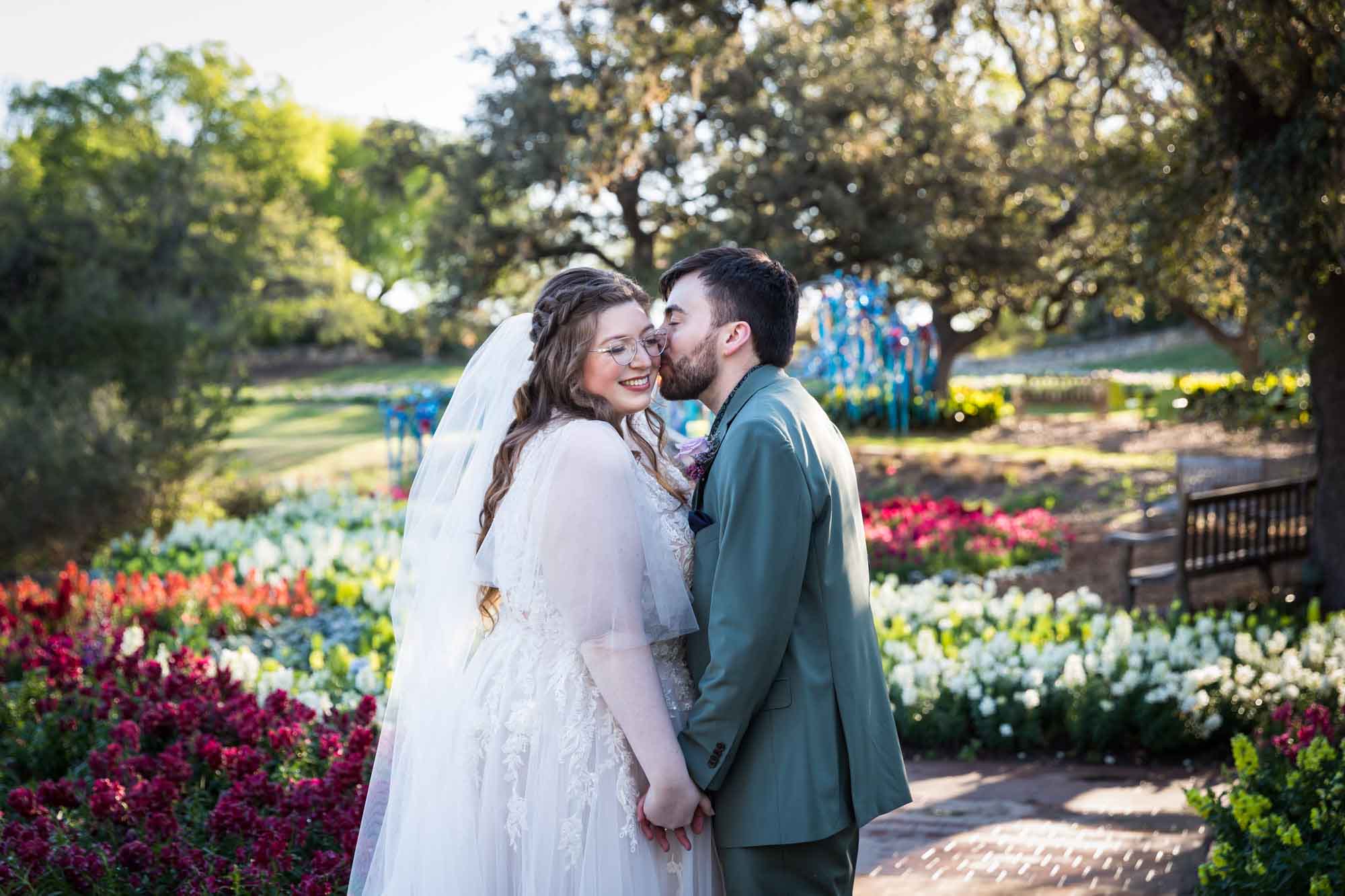 Groom kissing bride on the cheek and holding hands in front of bed of colorful snapdragons and trees before a San Antonio Botanical Garden wedding ceremony