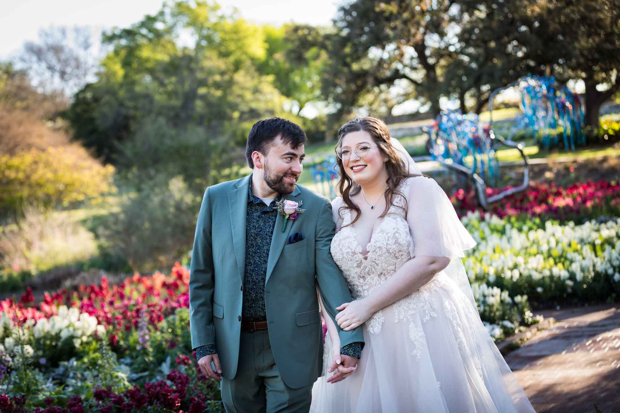 Bride and groom holding hands in front of bed of colorful snapdragons and trees before a San Antonio Botanical Garden wedding ceremony