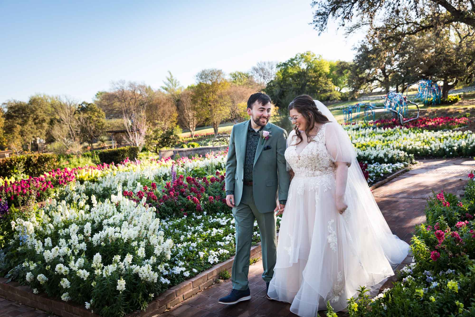 Bride and groom holding hands walking in front of bed of colorful snapdragons and trees before a San Antonio Botanical Garden wedding ceremony