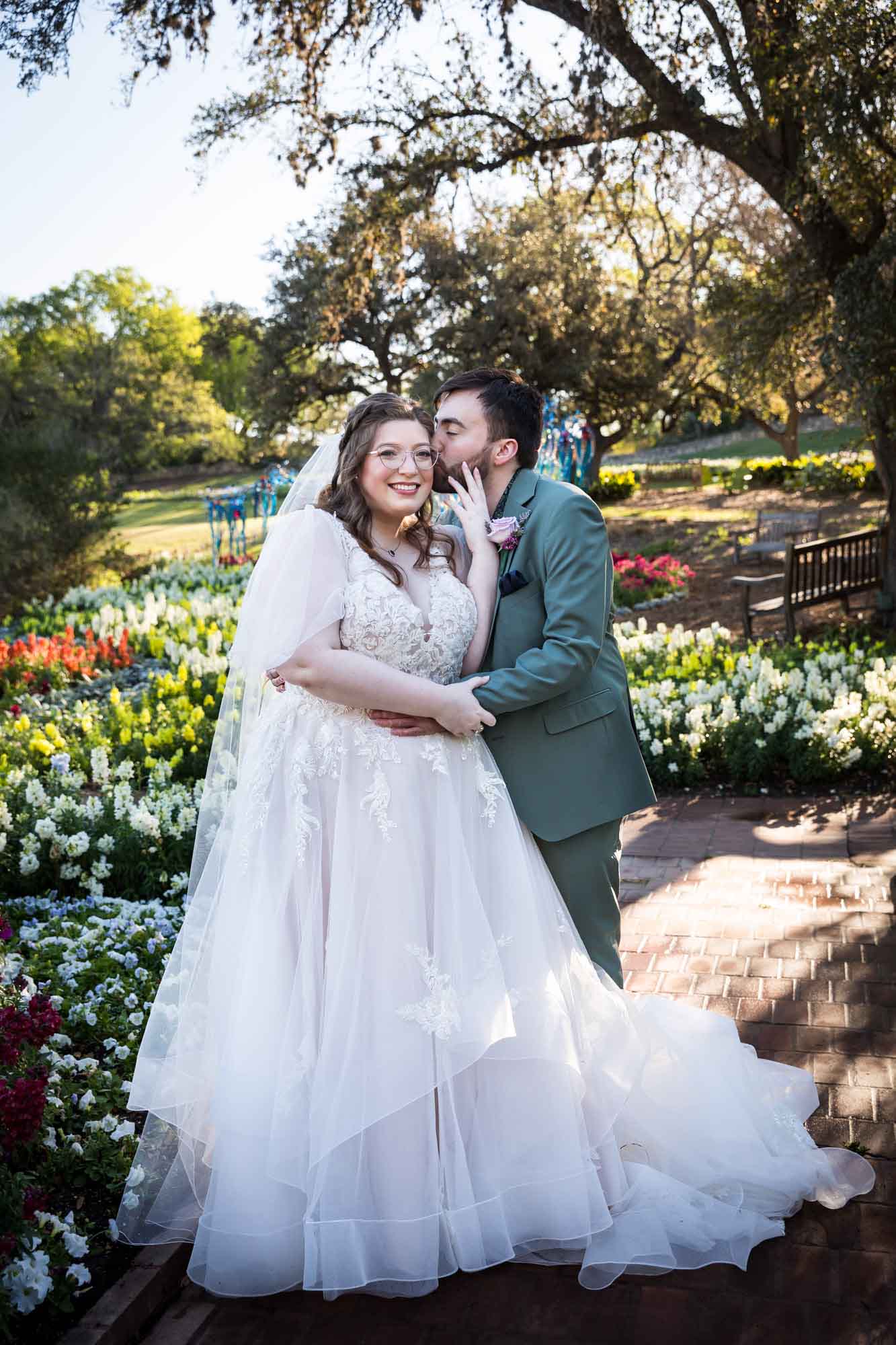 Groom kissing bride on side of head and hugging in front of bed of colorful snapdragons and trees before a San Antonio Botanical Garden wedding ceremony