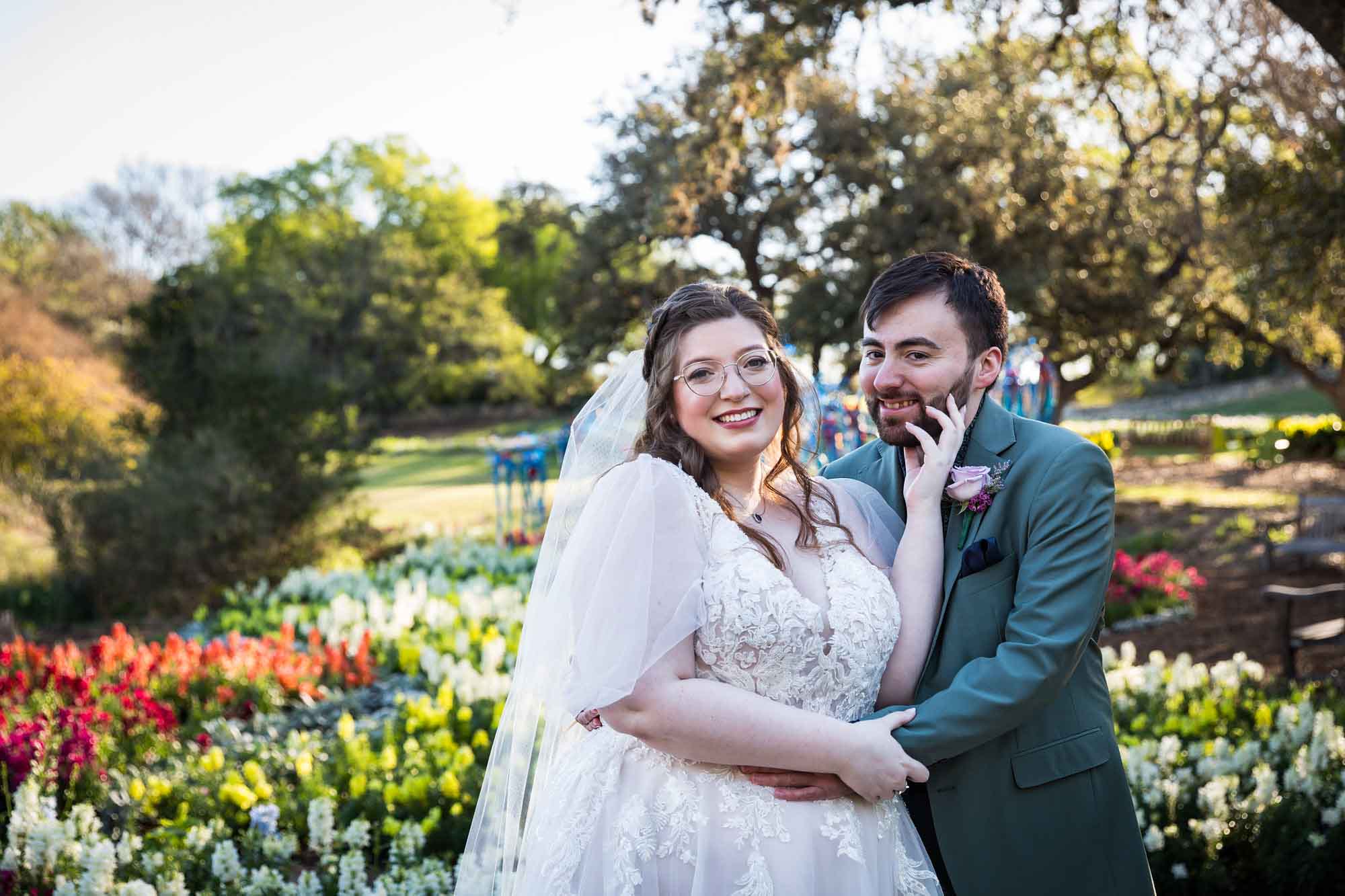 Bride and groom hugging in front of bed of colorful snapdragons and trees before a San Antonio Botanical Garden wedding ceremony