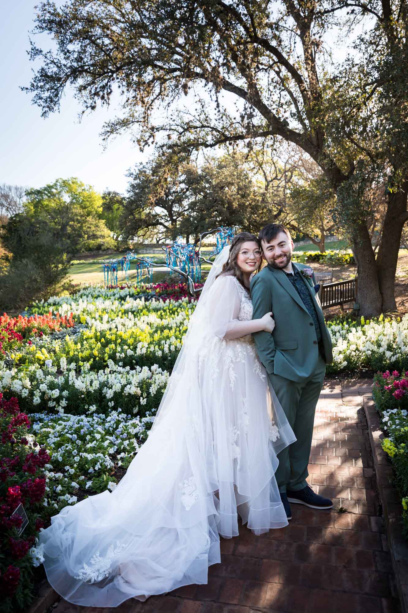 Bride and groom hugging in front of bed of colorful snapdragons and trees before a San Antonio Botanical Garden wedding ceremony