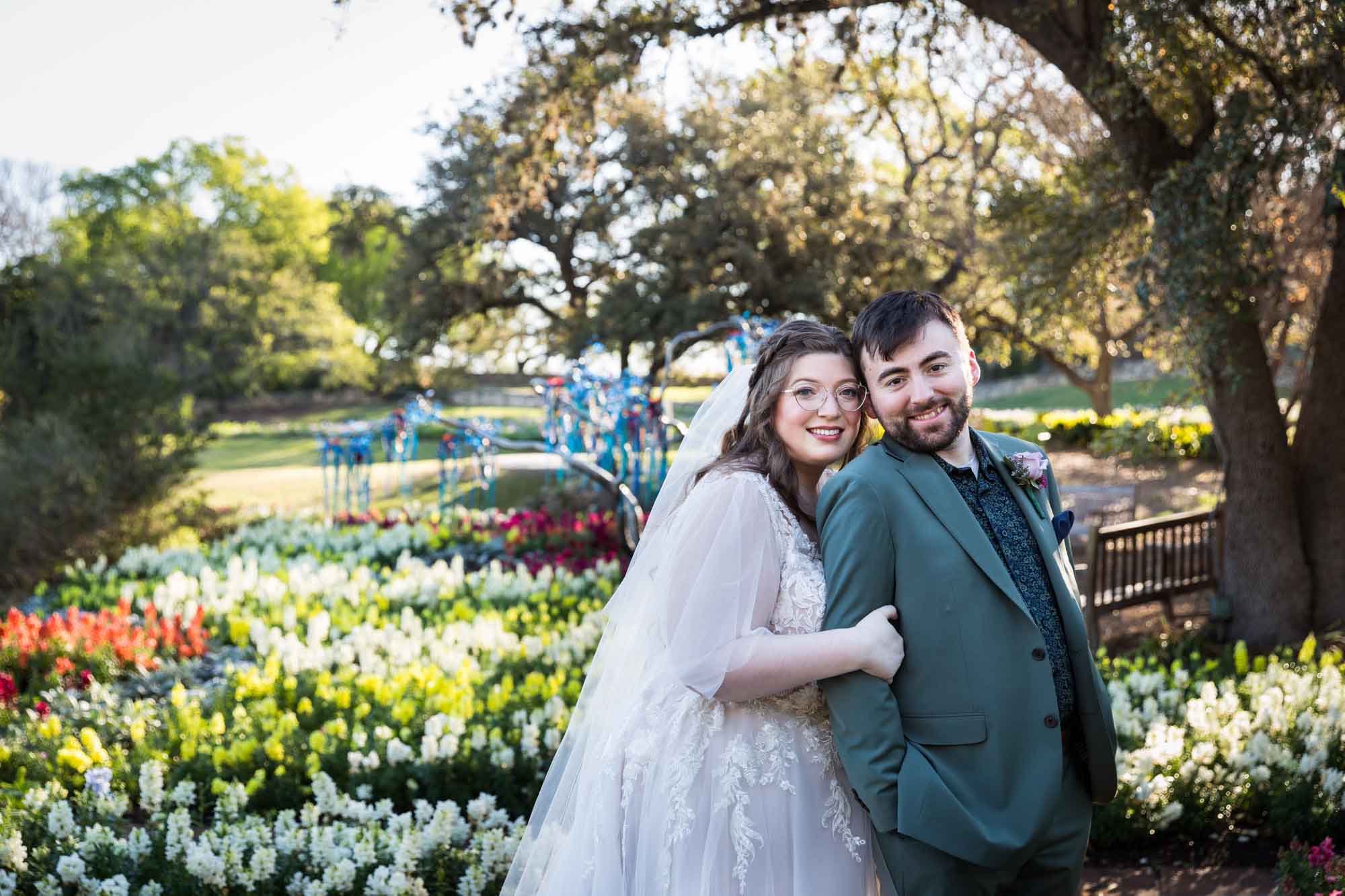 Bride and groom hugging in front of bed of colorful snapdragons and trees before a San Antonio Botanical Garden wedding ceremony