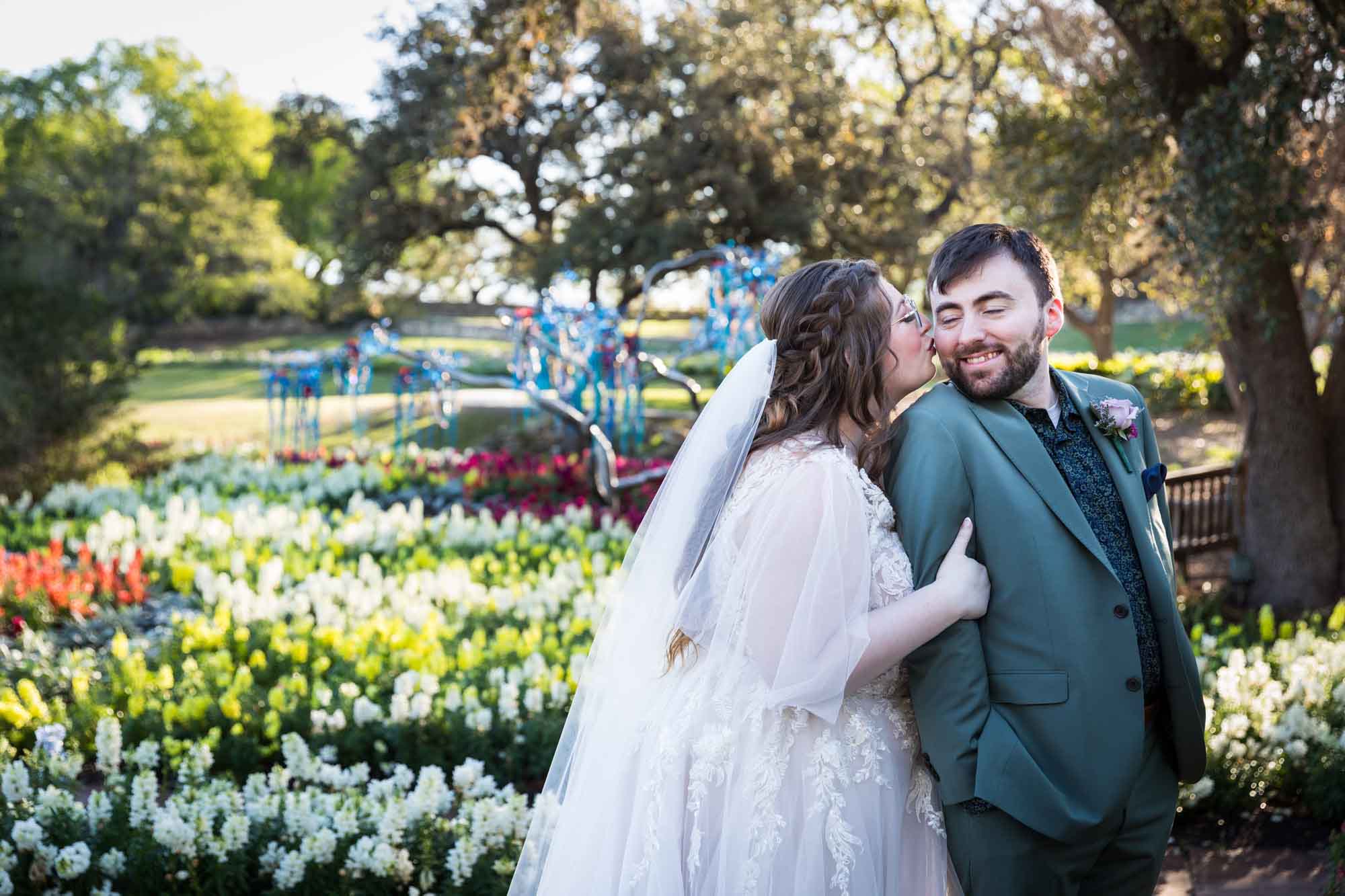 Bride kissing groom on cheek in front of bed of colorful snapdragons and trees before a San Antonio Botanical Garden wedding ceremony