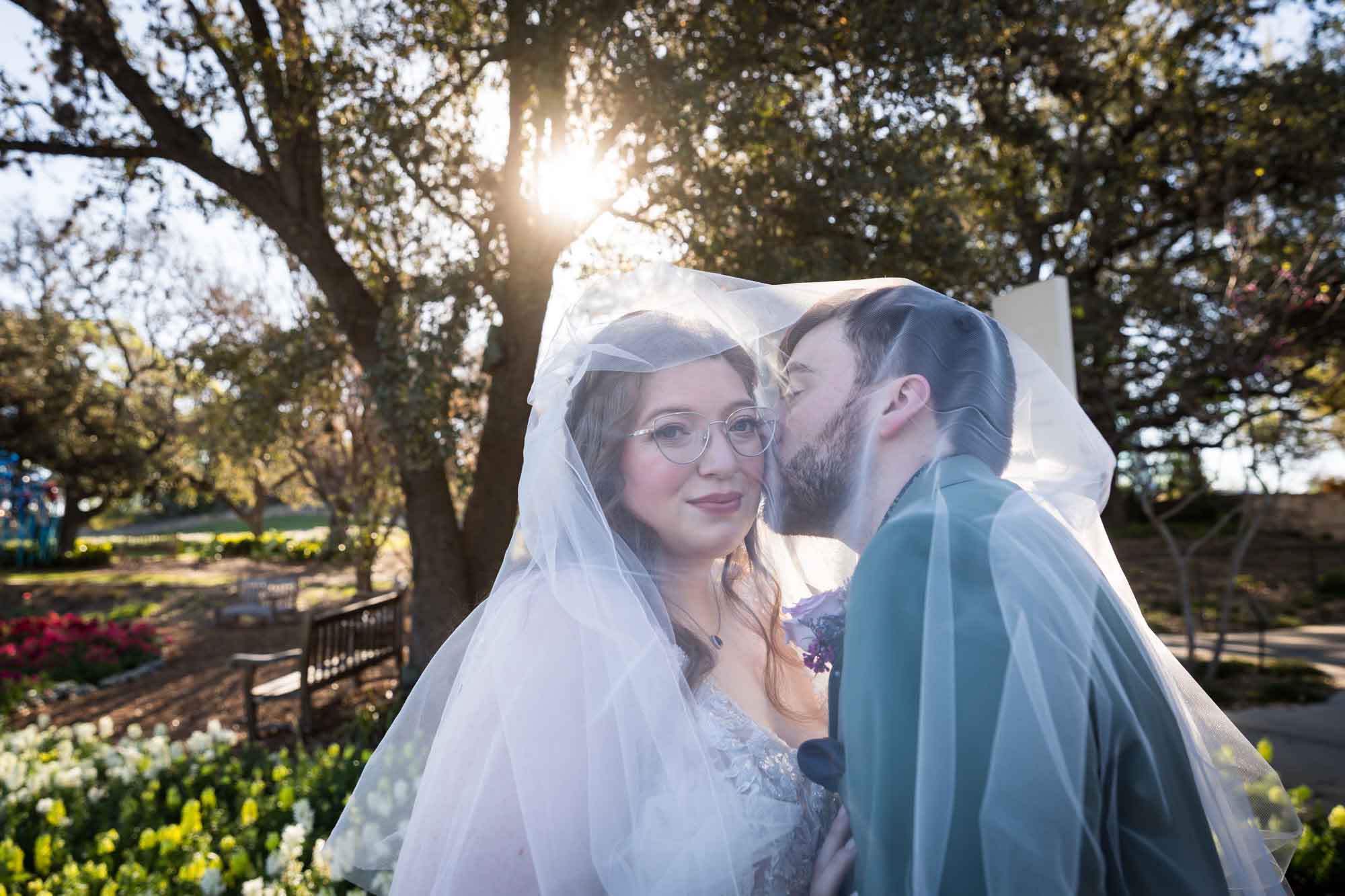 Groom kissing bride on the cheek underneath veil in front of tree before a San Antonio Botanical Garden wedding ceremony