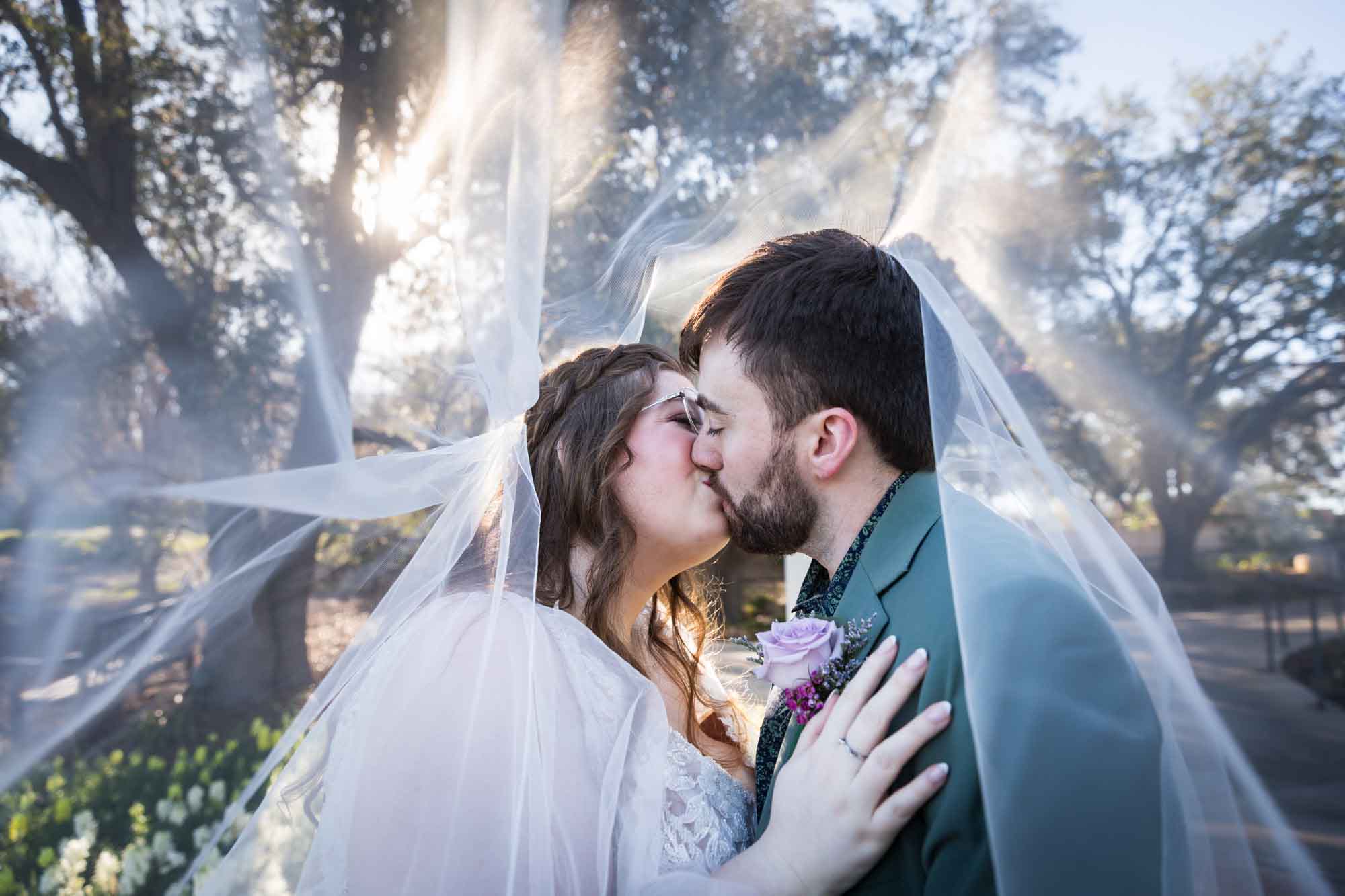 Bride and groom kissing underneath a veil in front of trees before a San Antonio Botanical Garden wedding ceremony
