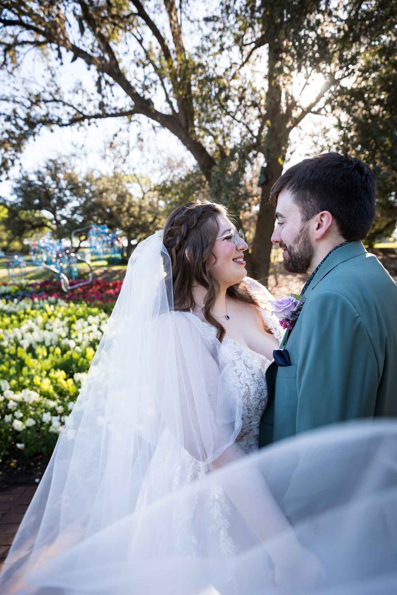 Bride and groom standing behind veil and in front of colorful flowers and trees before a San Antonio Botanical Garden wedding ceremony