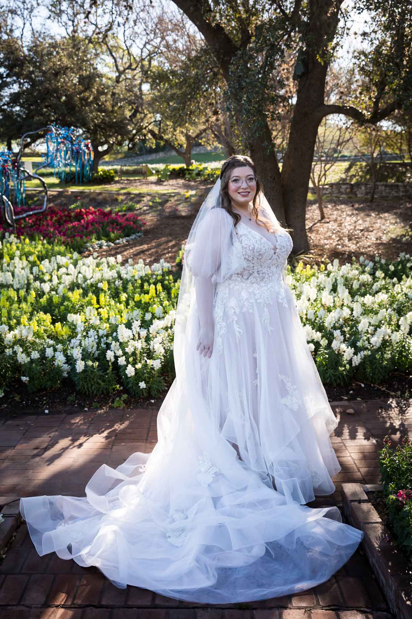 Bride wearing white dress and veil in front of colorful flowers and tree before a San Antonio Botanical Garden wedding ceremony