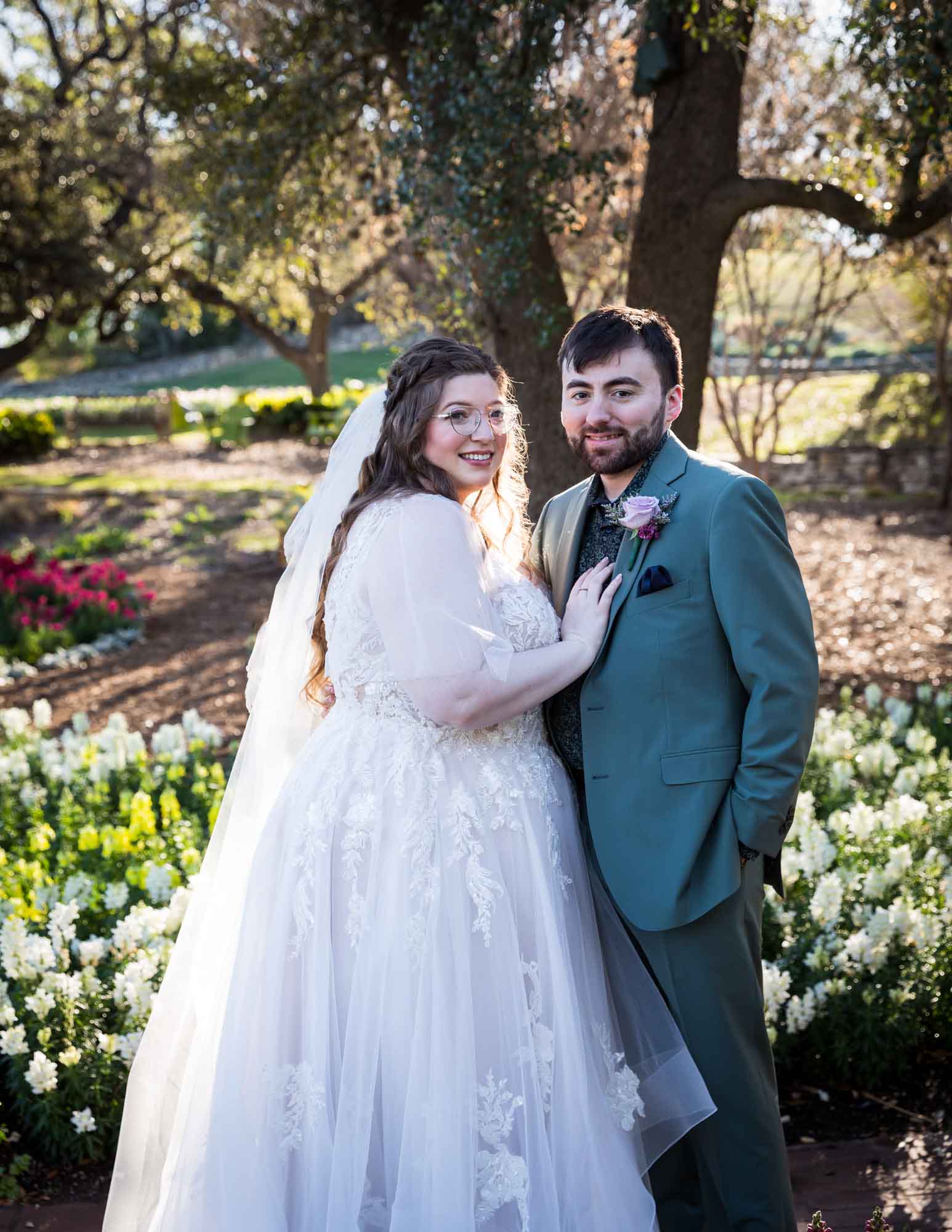 Bride standing beside groom with hand on his chest in front of colorful flowers and tree before a San Antonio Botanical Garden wedding ceremony