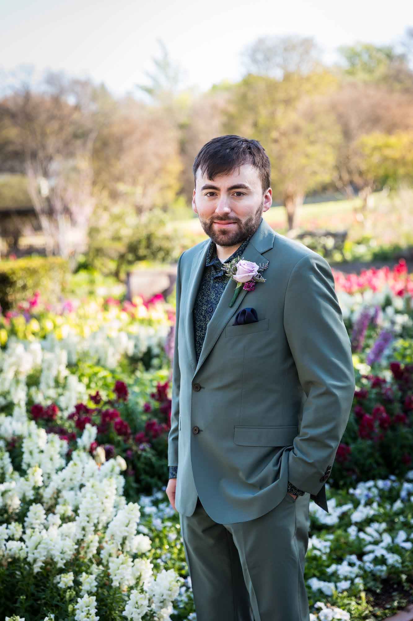 Groom wearing green suit standing with hands in pockets in front of colorful flowers and trees before a San Antonio Botanical Garden wedding ceremony