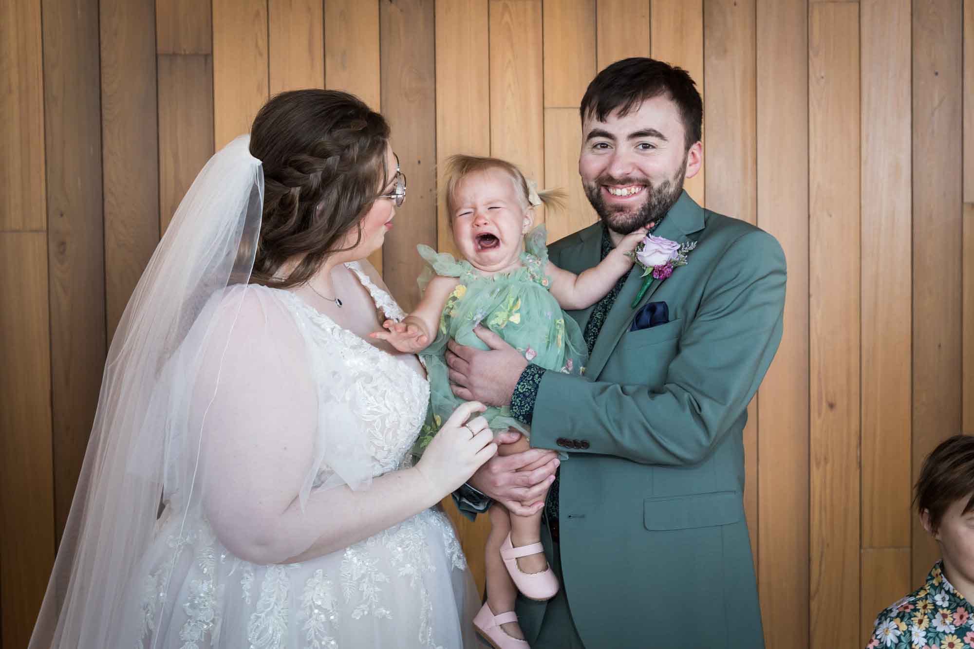 Crying baby being held by groom wearing green suit beside bride before a San Antonio Botanical Garden wedding ceremony