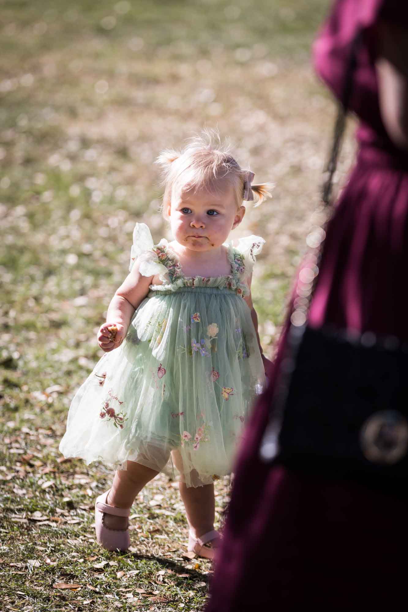 Little girl wearing green dress with crumbs on her mouth walking in grass before a San Antonio Botanical Garden wedding ceremony
