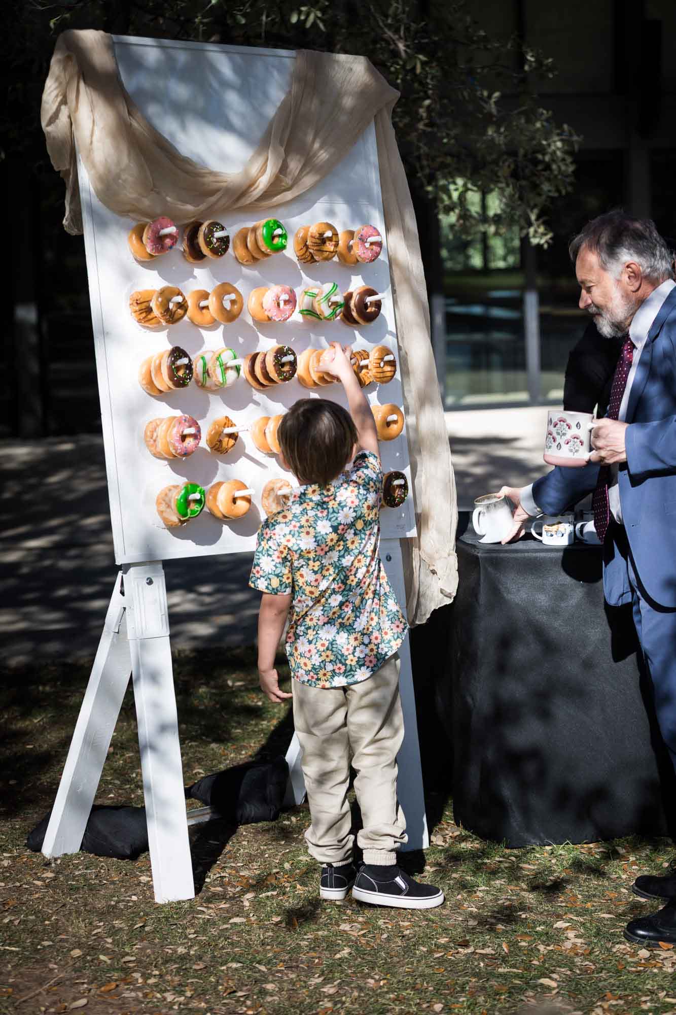 Little boy reaching up for doughnut hanging from doughnut wall outdoors before a San Antonio Botanical Garden wedding ceremony