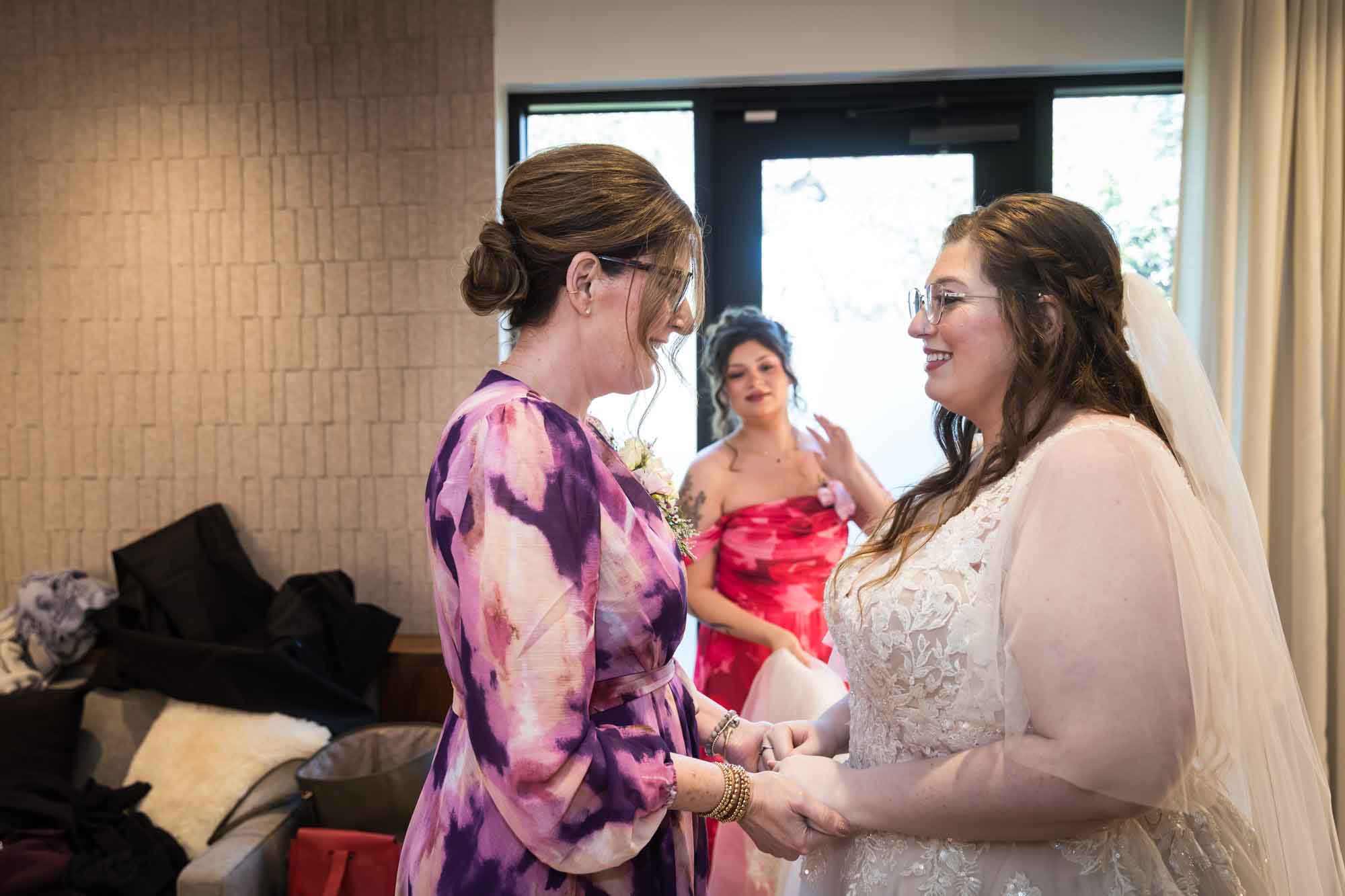 Bride holding hands with mother wearing purple floral dress with woman wearing red floral dress in the background before a San Antonio Botanical Garden wedding ceremony